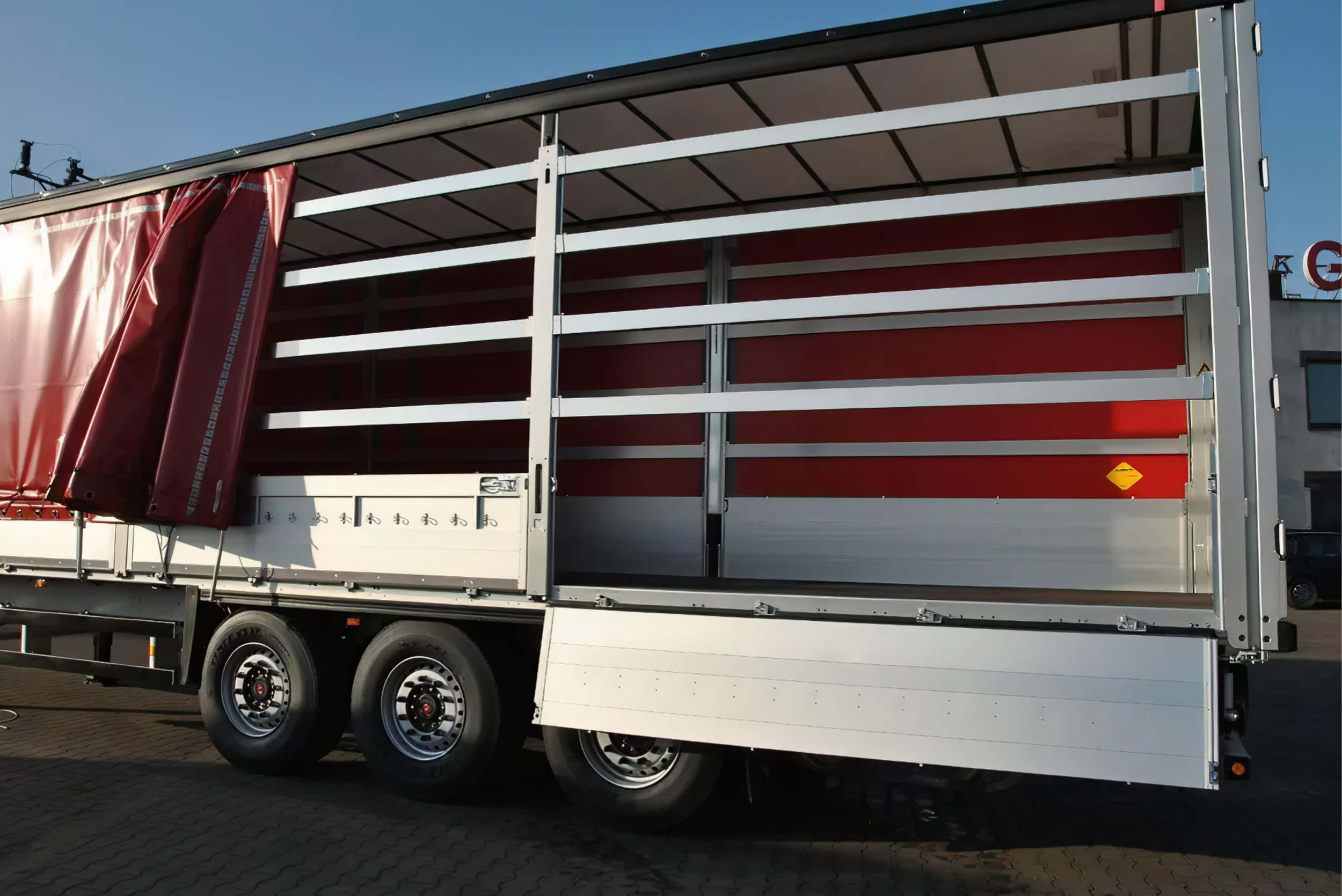 A large flatbed truck with a red tarpaulin cover partially pulled back, parked on a paved area under a clear blue sky.