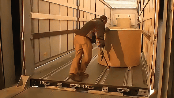 A person loading a large cardboard box into a truck, with wood-paneled walls and a metal floor.