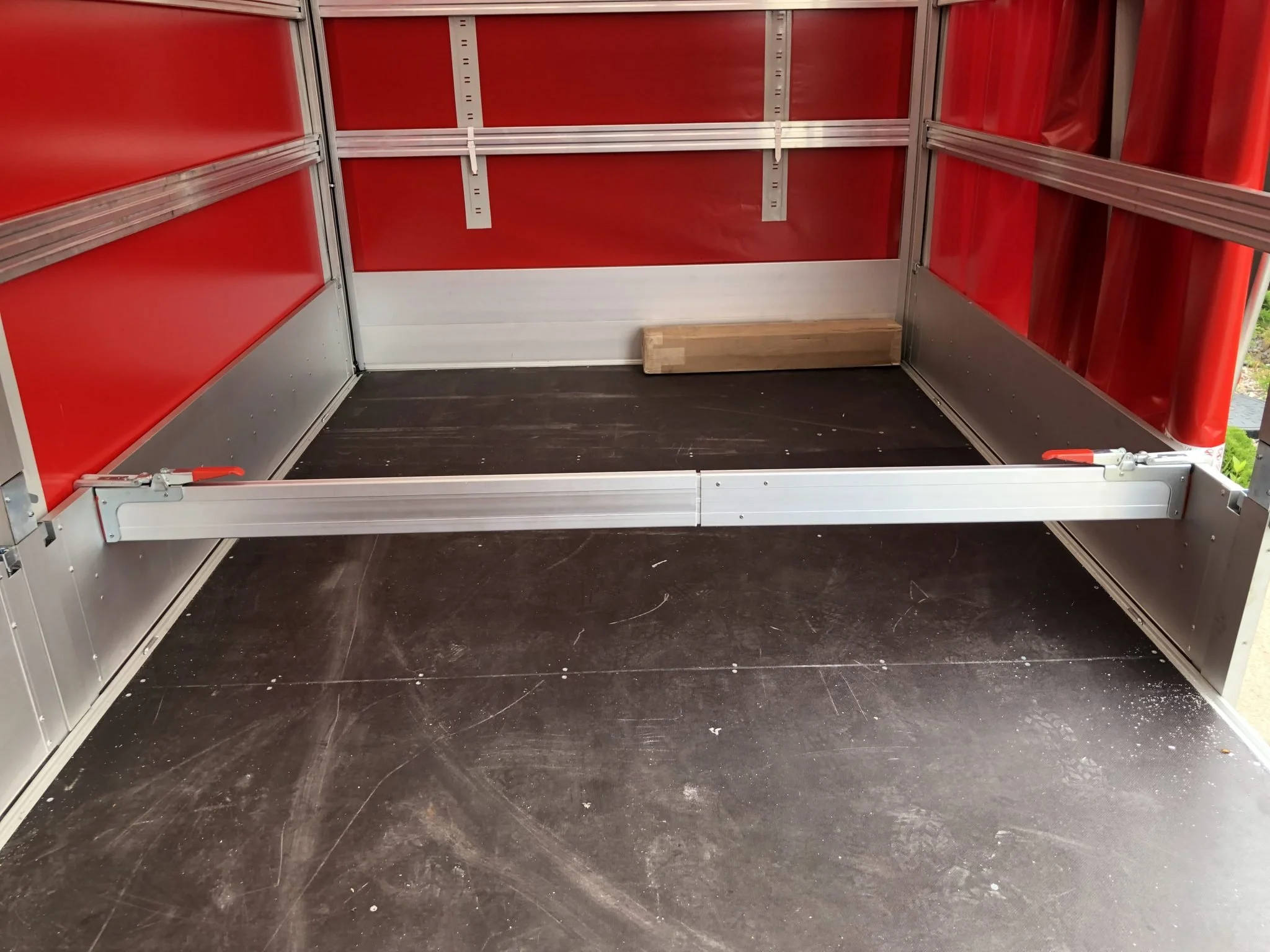 Empty retail display shelf with red plastic lining, metal framing, and a wooden block on the dark floor.