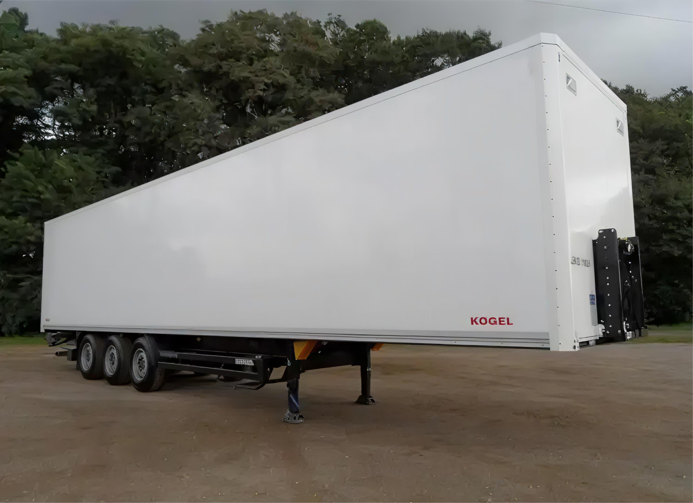 A large white semi-trailer truck with the brand name 'KOGEL' written on the side, parked on a dirt surface with a background of dense green trees.