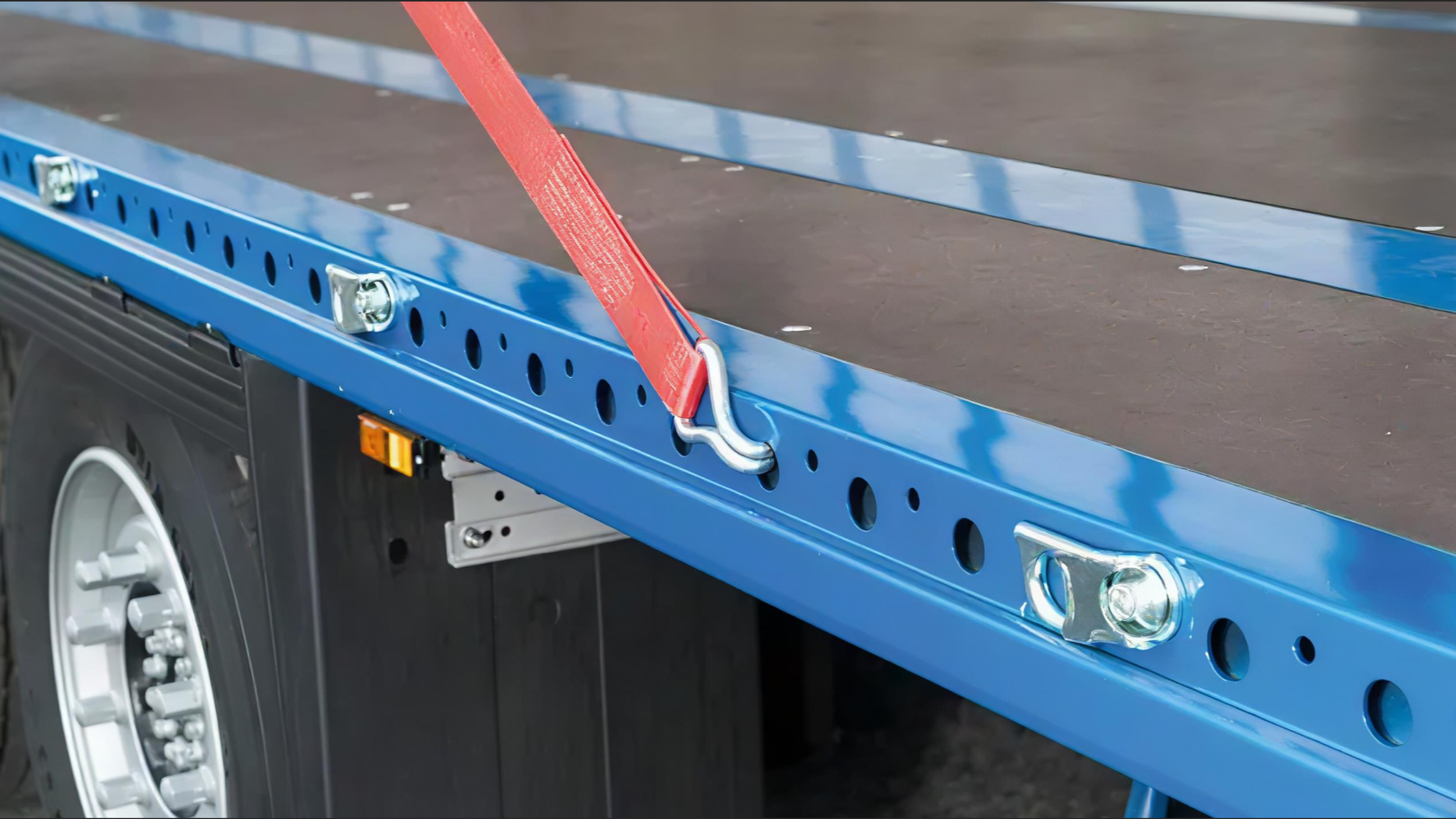 Close-up of a blue flatbed truck with a red strap securing a load, showing part of the truck's wheel and metallic framework.