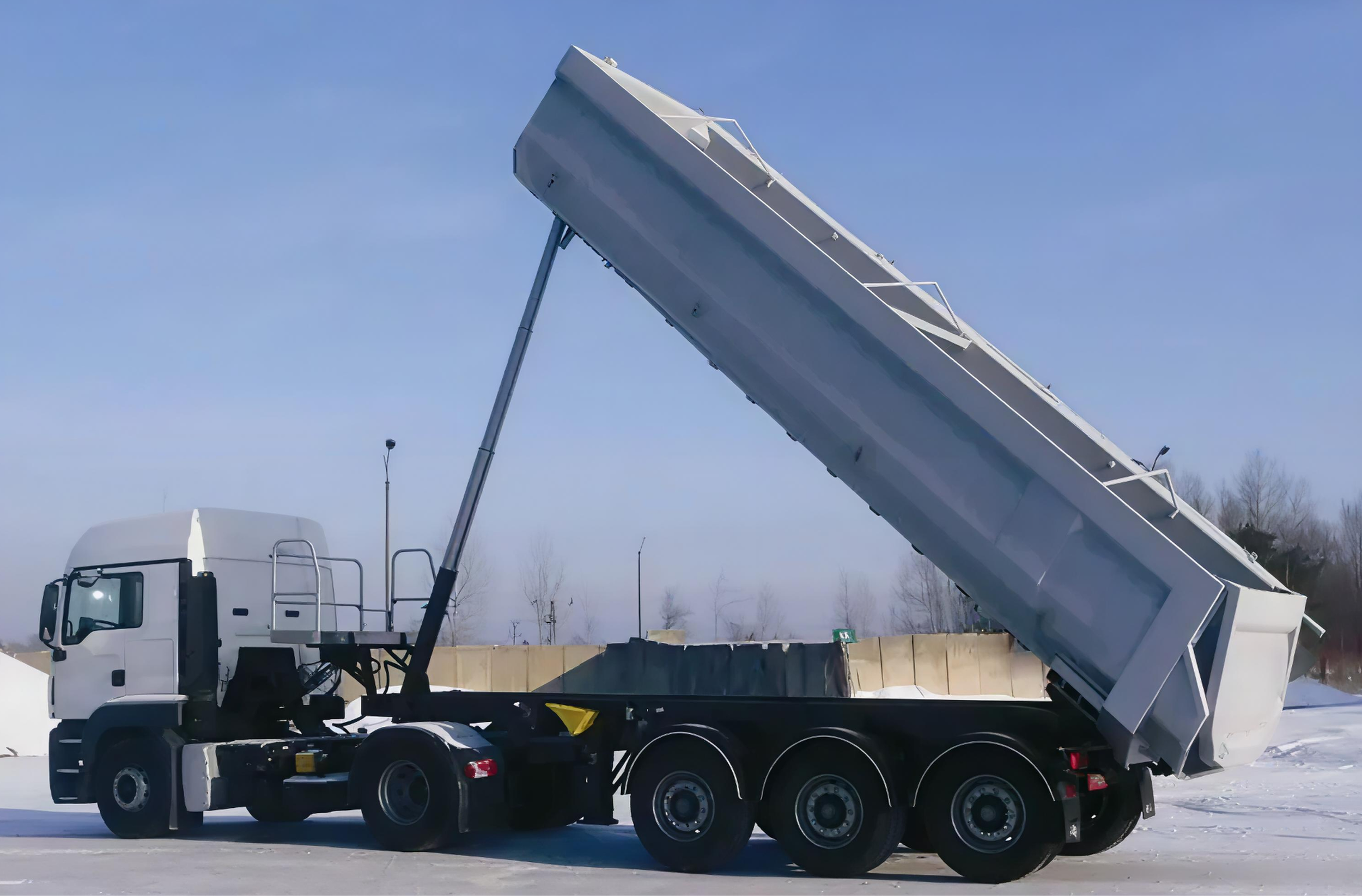 A white truck with a large gravel or cement mixer drum on the back, tilted upward, parked on snow-covered ground with a clear blue sky and a few trees in the background.