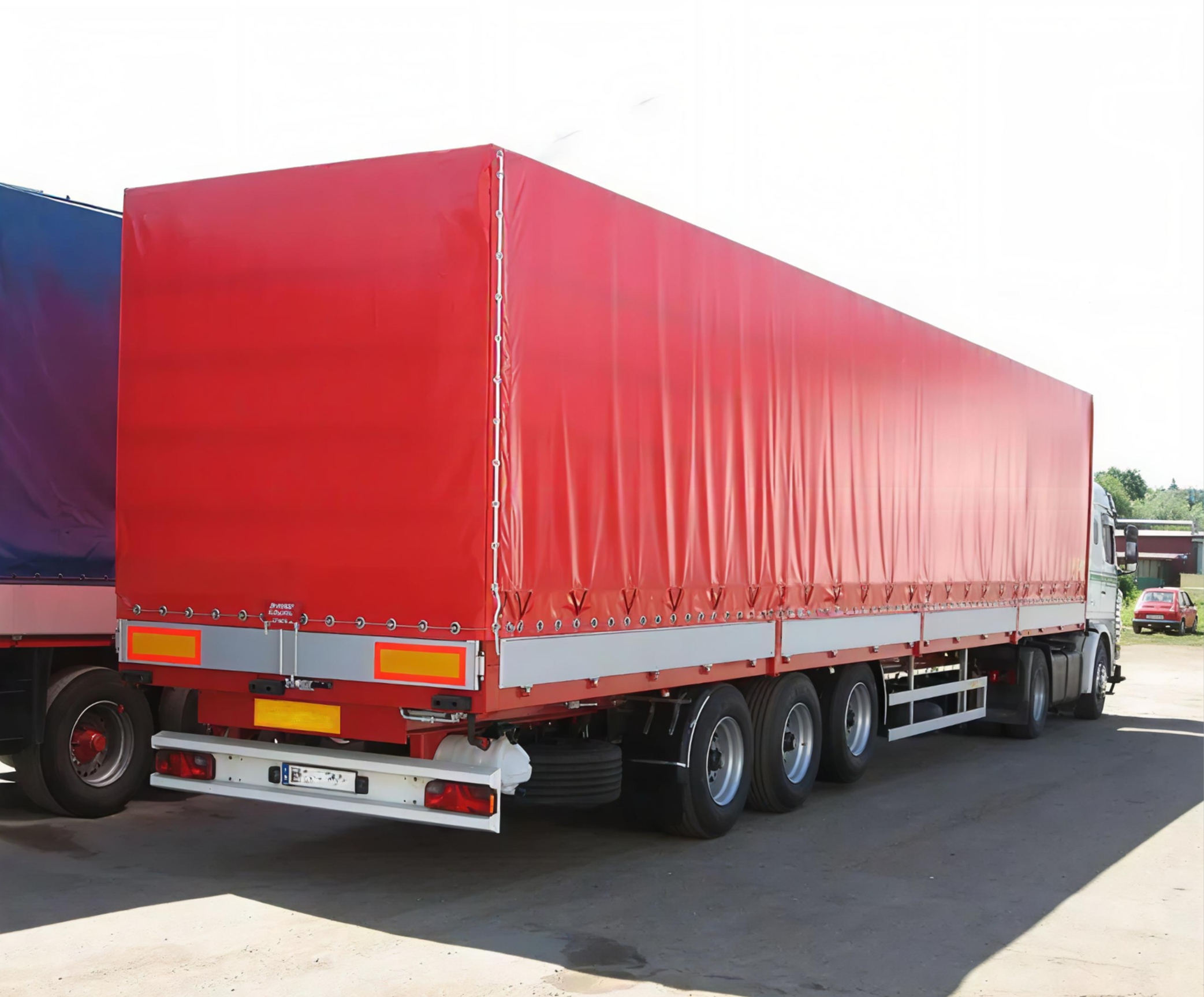 A red cargo truck with a closed, red trailer parked outdoors on a paved surface, with some greenery and a blue vehicle visible in the background.