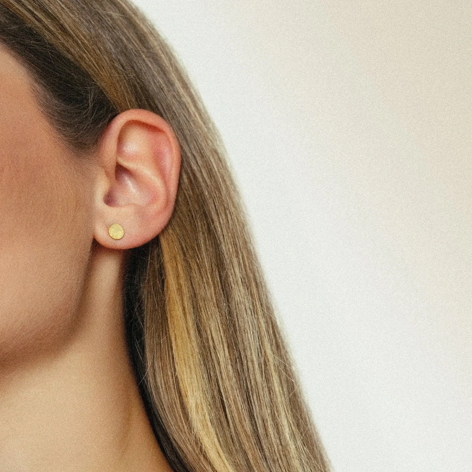 Nahaufnahme eines Ohrläppchens mit kleinem goldfarbenem Stecker, glatte Haut, braune Haare, heller Hintergrund.