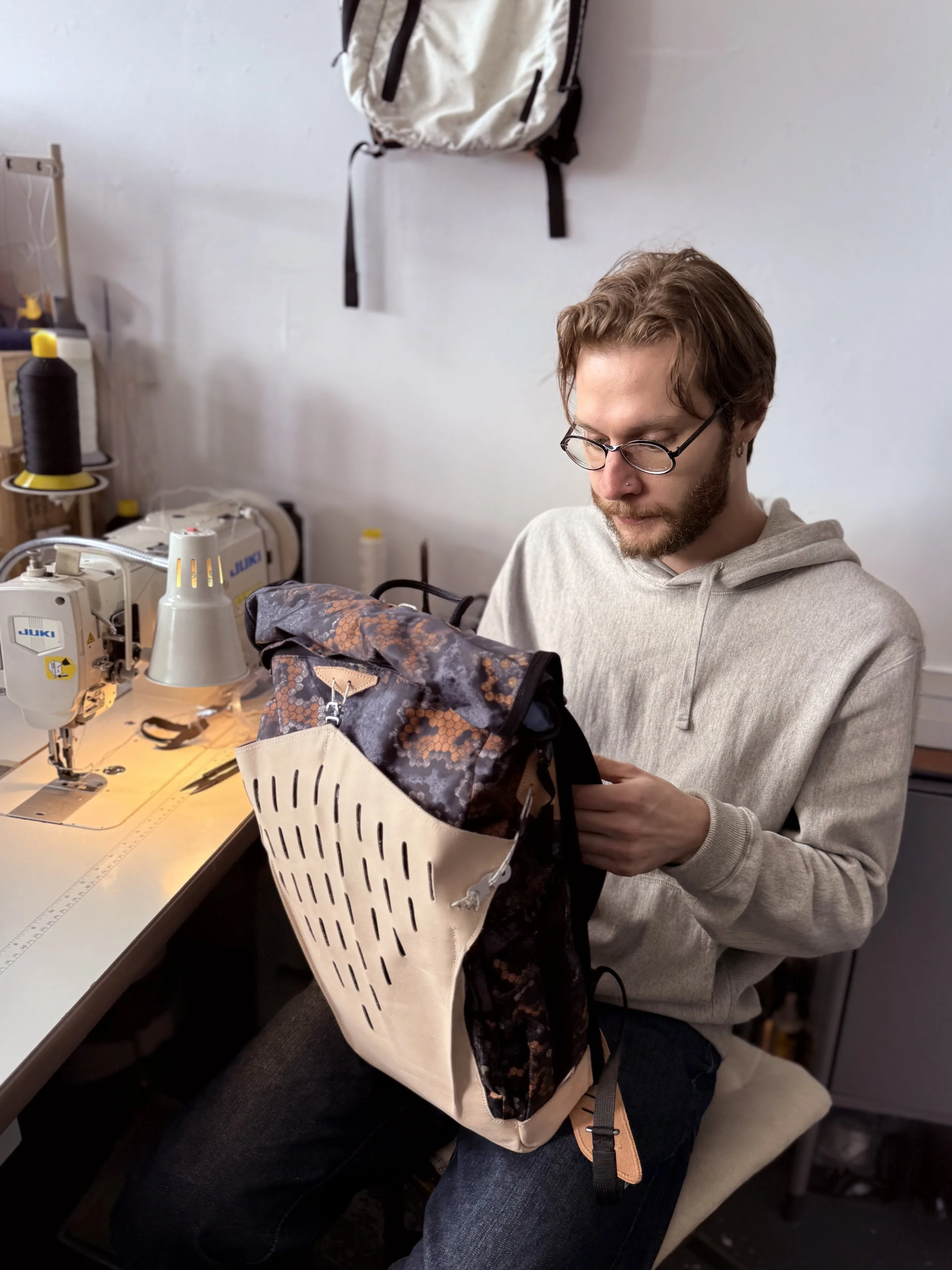 A man with glasses and a beard, wearing a light gray hoodie, sewing in a workshop. He is working with fabric on a sewing machine covered with camouflage fabric, with a white plastic basket holding some of the fabric. A backpack is hanging on the wall behind him.