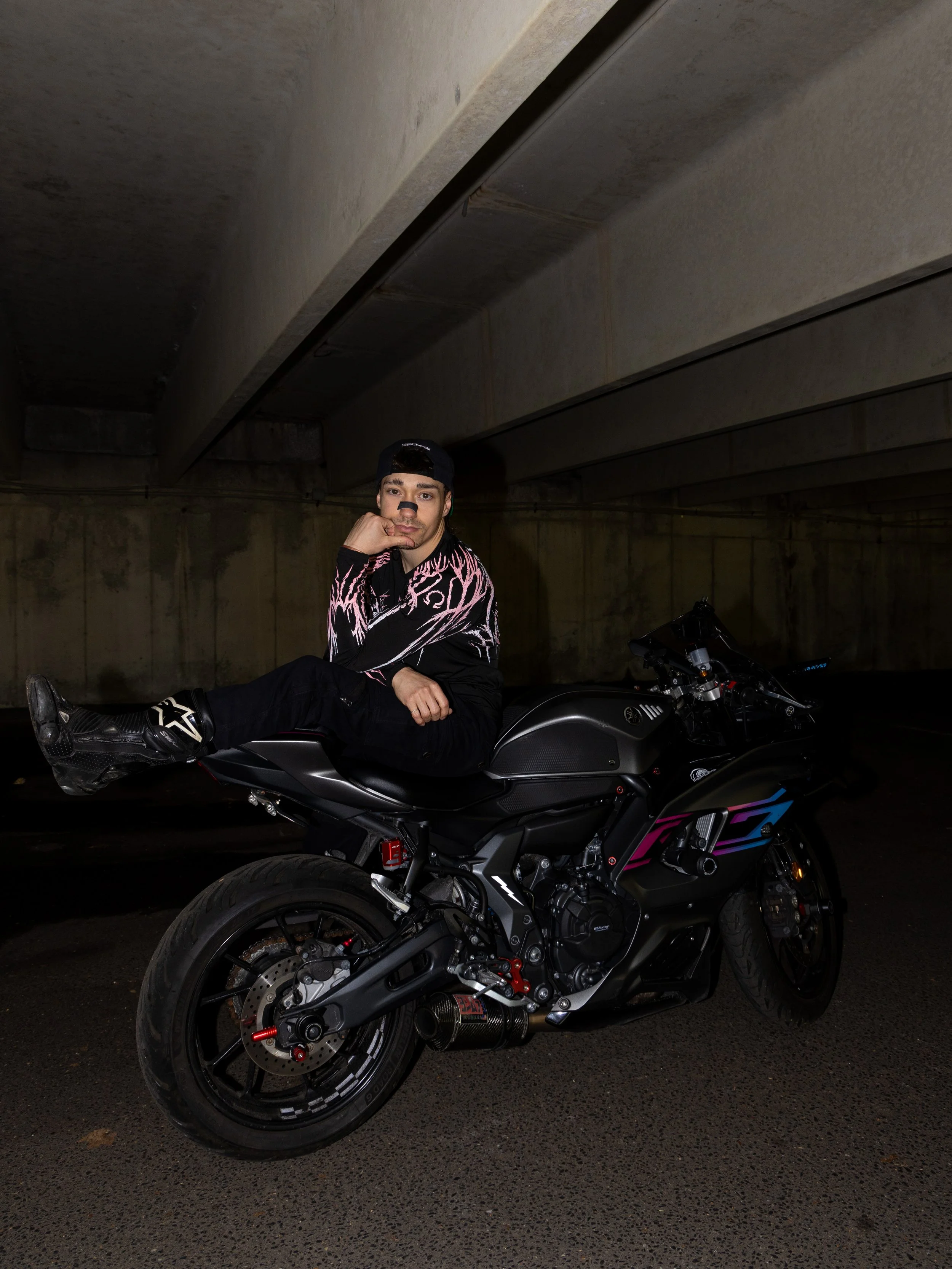 A young man sitting on a black motorcycle in a dimly lit parking garage. He is wearing a black cap backwards, a black and pink patterned jacket, and black pants. The motorcycle is parked on a textured asphalt surface.