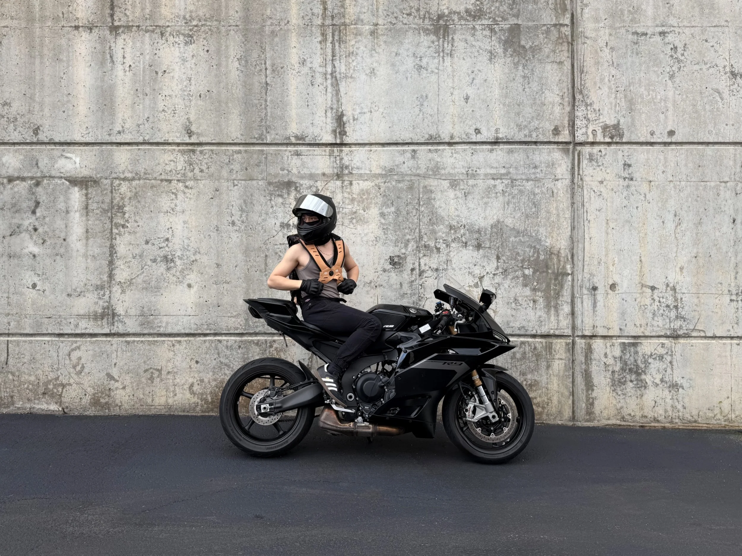 Person sitting on a black motorcycle, wearing a helmet, gloves, and a vest, in front of a concrete wall.