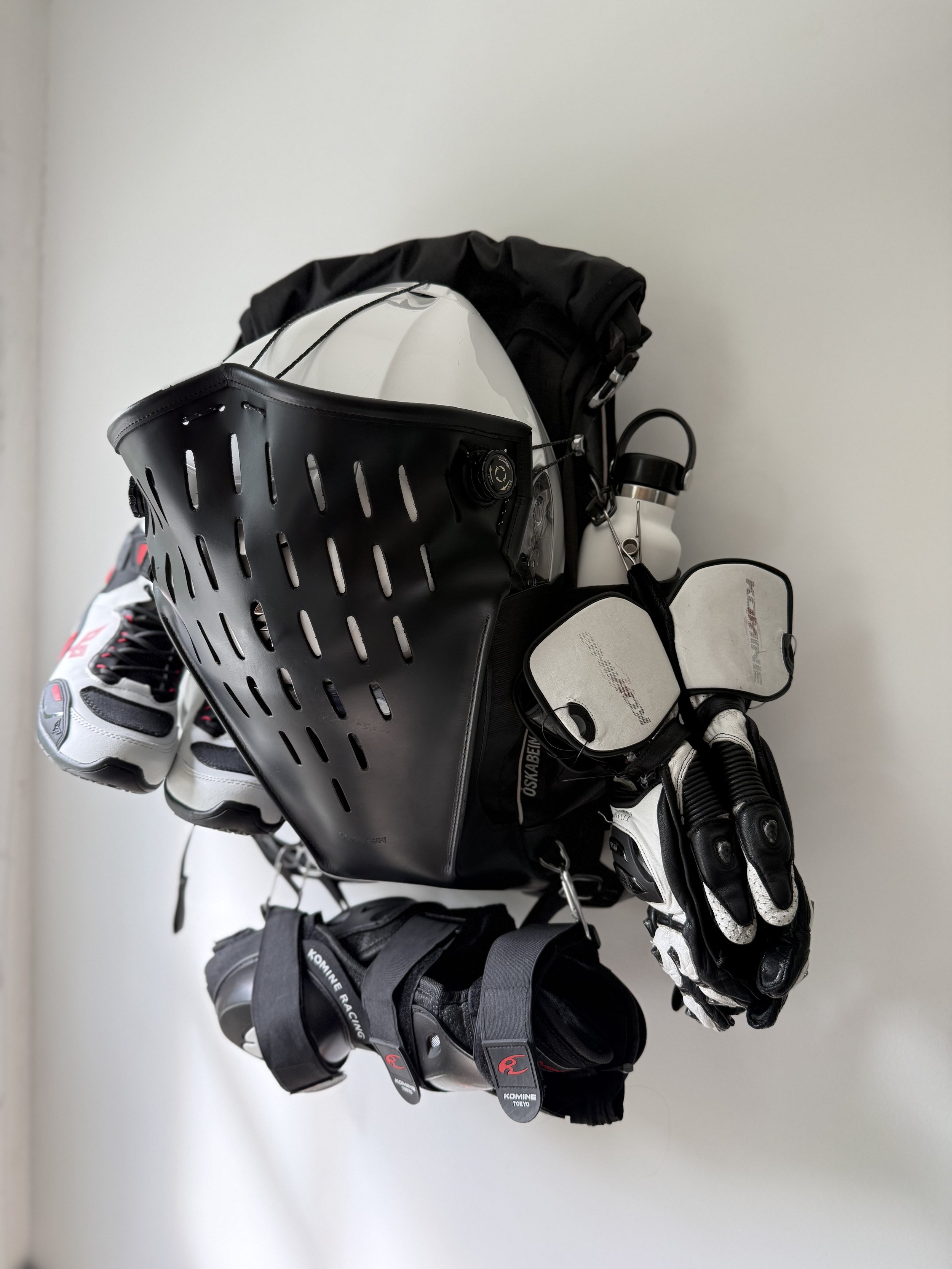 A collection of motorcycle equipment including riding shoes, gloves, knee guards, and a water bottle, attached to a backpack.