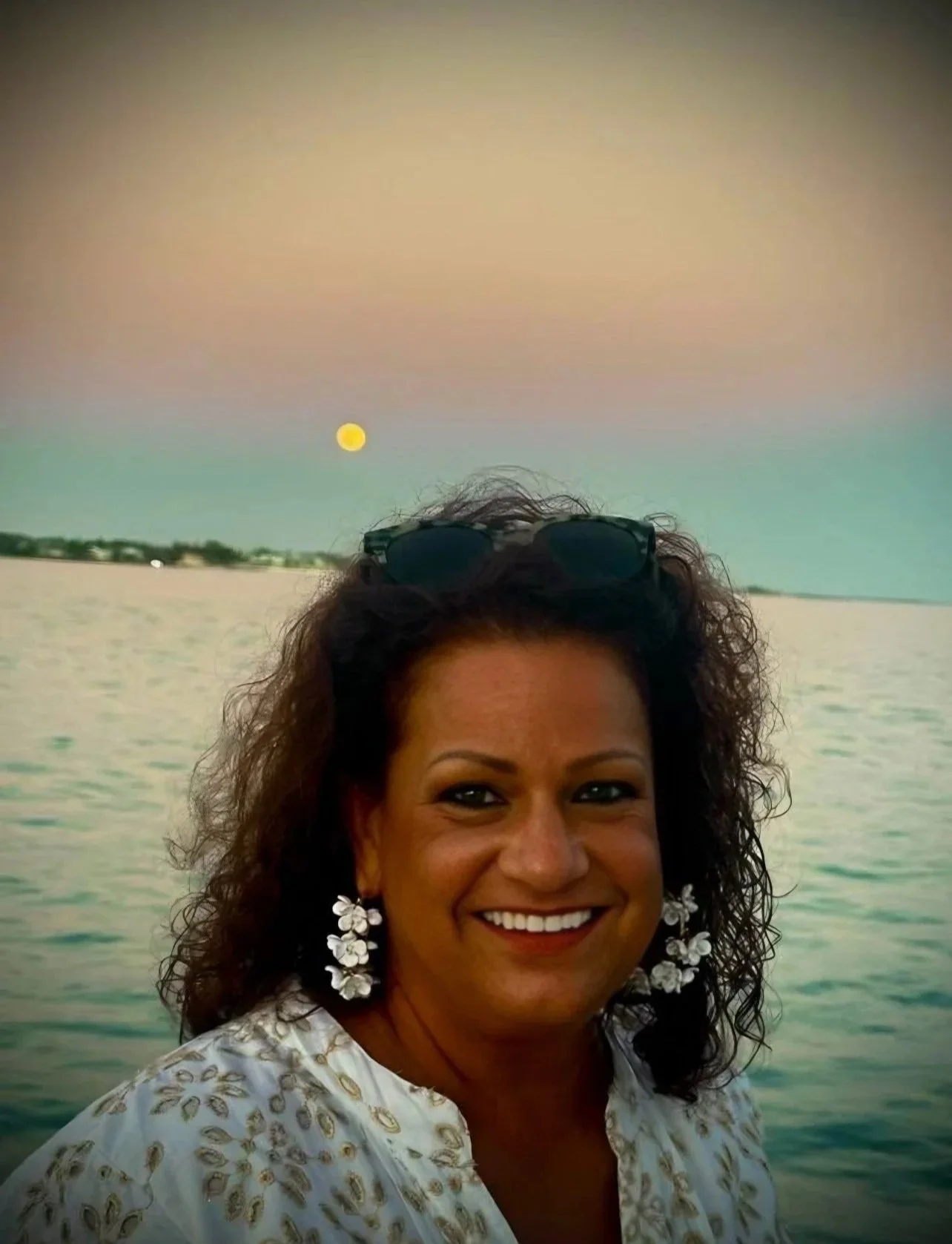 A woman smiling at the camera with a body of water and a full moon in the background during sunset or dusk.
