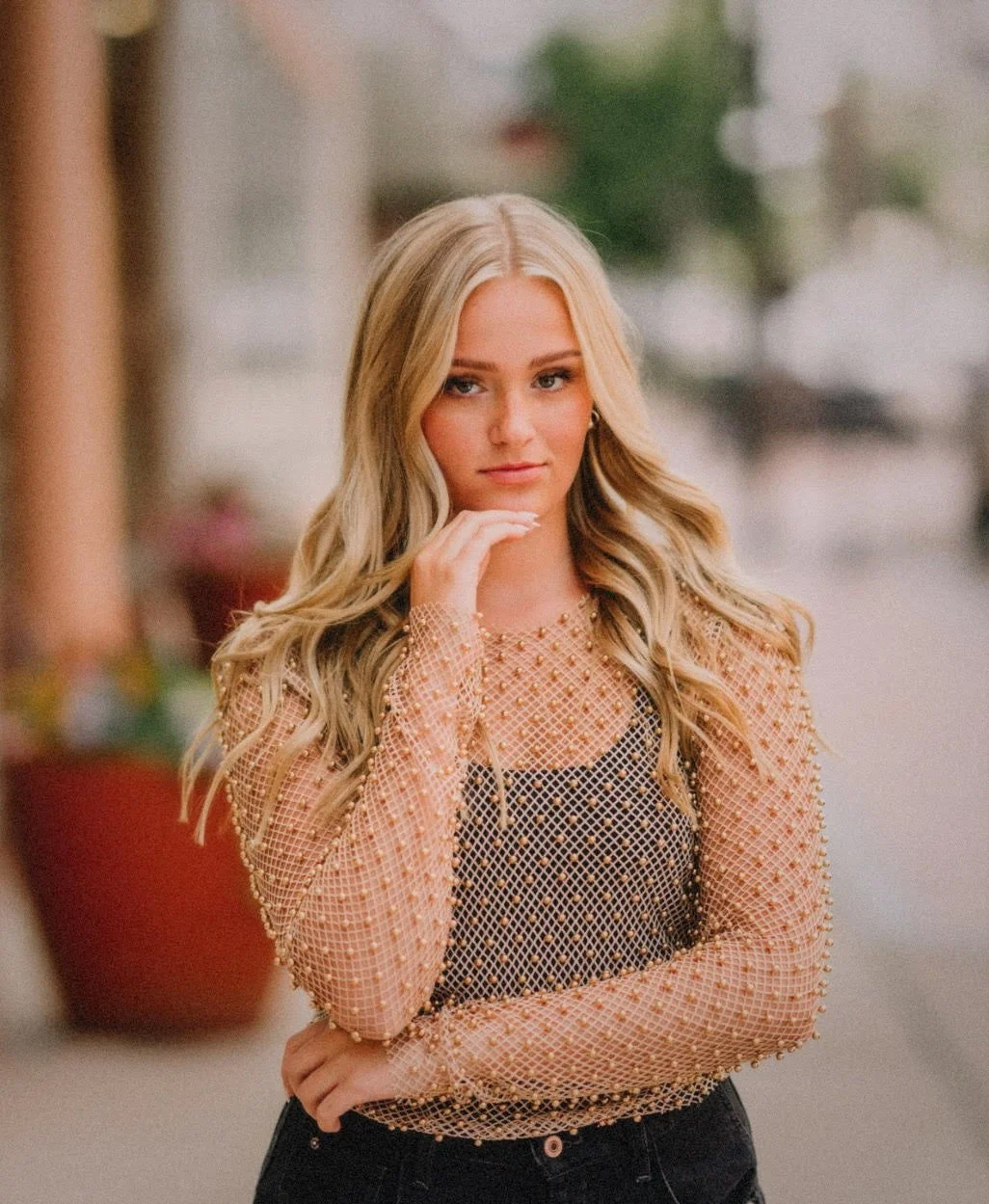 Young woman with blonde hair, wearing a black top and an embellished, semi-transparent peach long-sleeve shirt, looking at the camera with her hand resting on her chin, outdoors with blurred background.