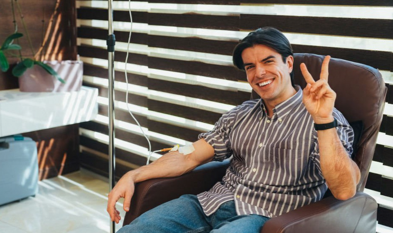 A man with dark hair smiling and making a peace sign while sitting in a leather chair in a room with a wooden slat wall. He has an IV in his arm and an IV pole next to him.