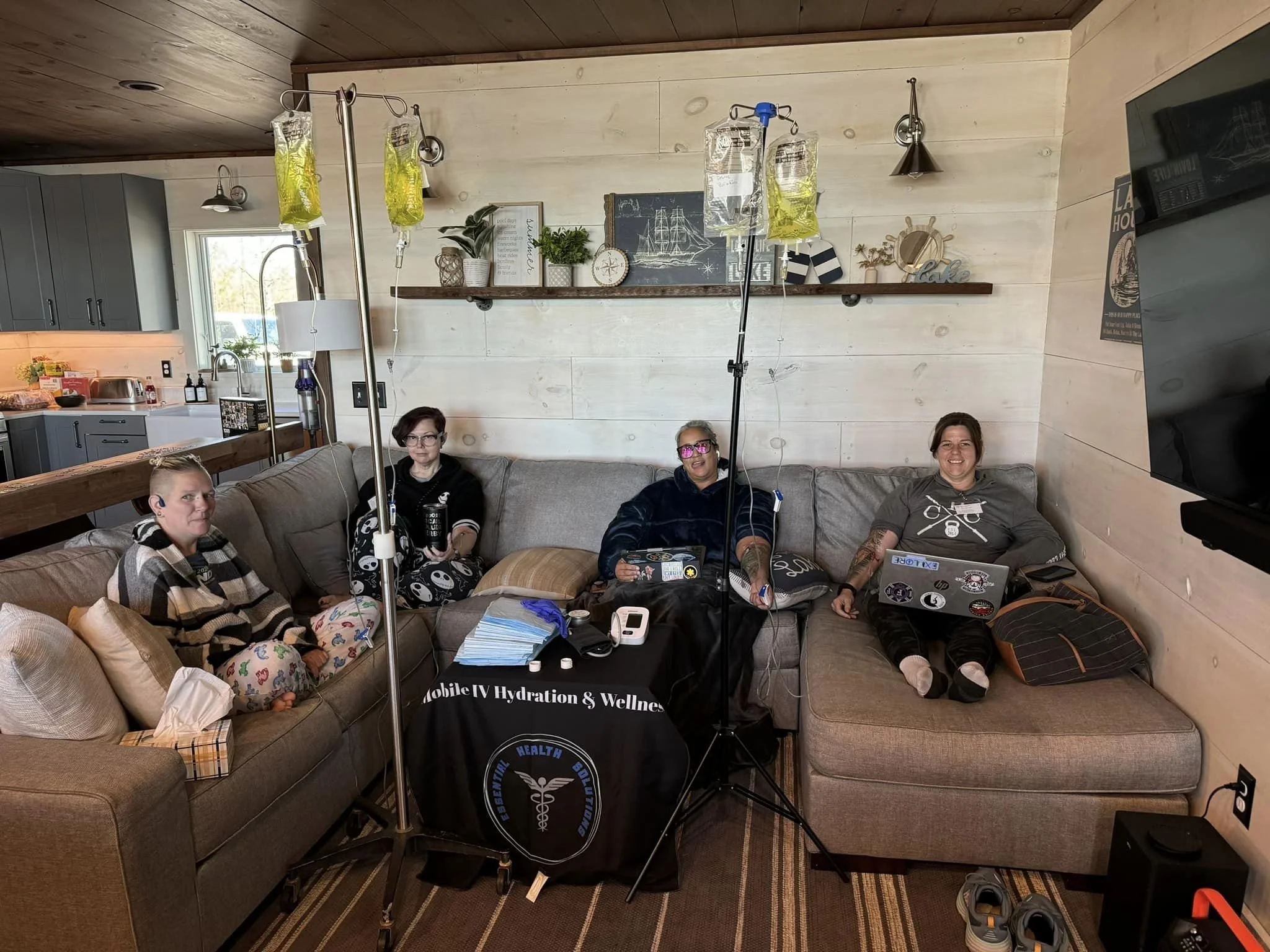Four women sitting on a beige sectional sofa inside a living room, with IV bags hanging from stands overhead, connected to one another. The women are smiling and using laptops, with medical supplies and equipment on a table in front of them. The living room has light-colored wooden walls, decor, and a television mounted on the wall.