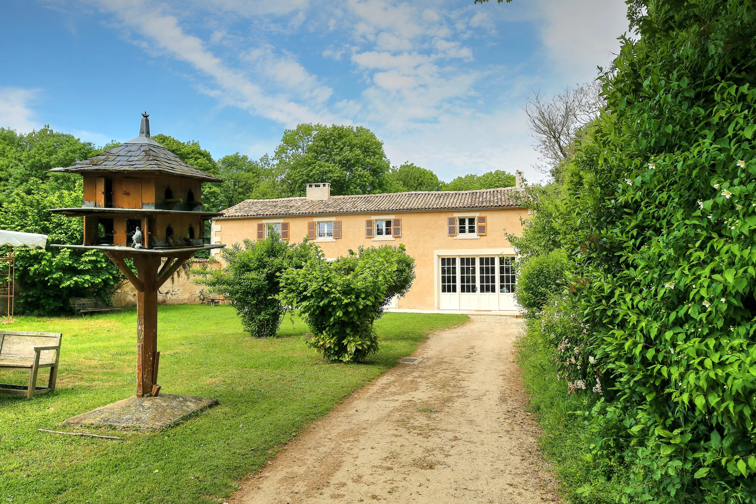 Jardin avec un chemin en terre, une maison beige avec des volets en bois, un phare en bois, et des arbres verts sous un ciel bleu avec quelques nuages.