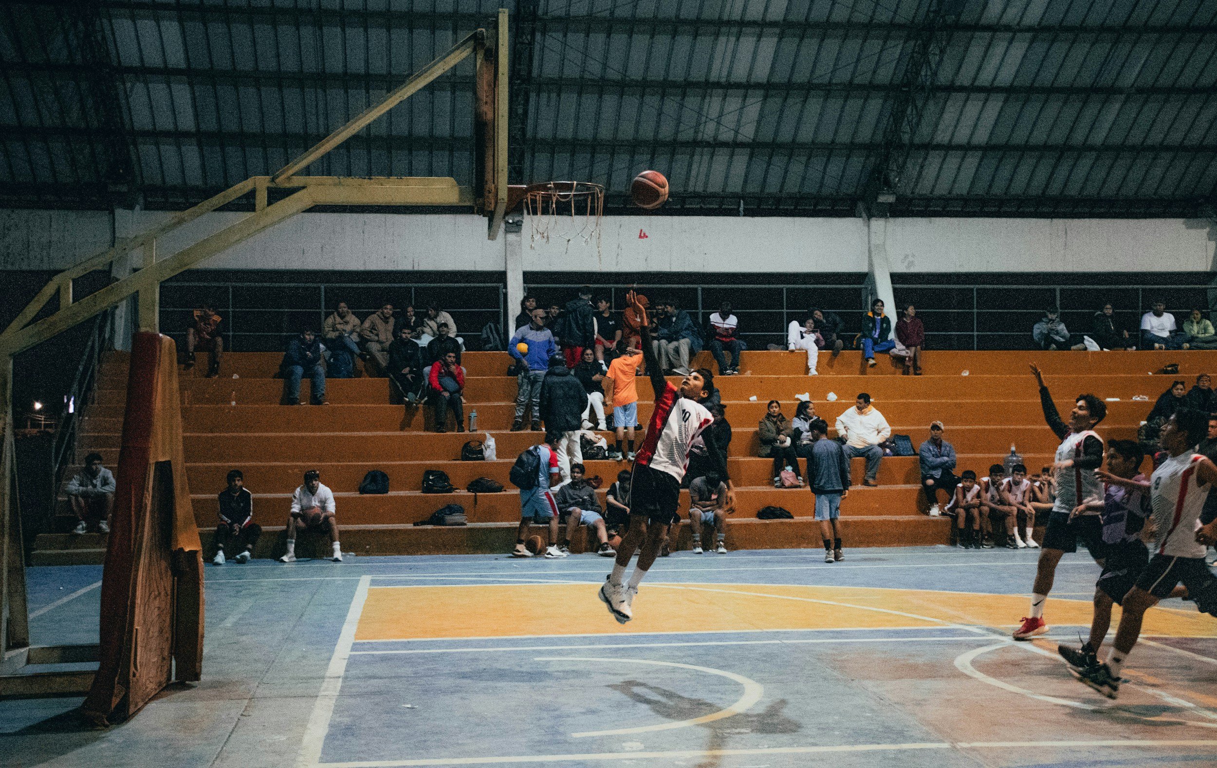 Indoor basketball game with players jumping to shoot or block the ball, spectators seated on orange benches in the background.