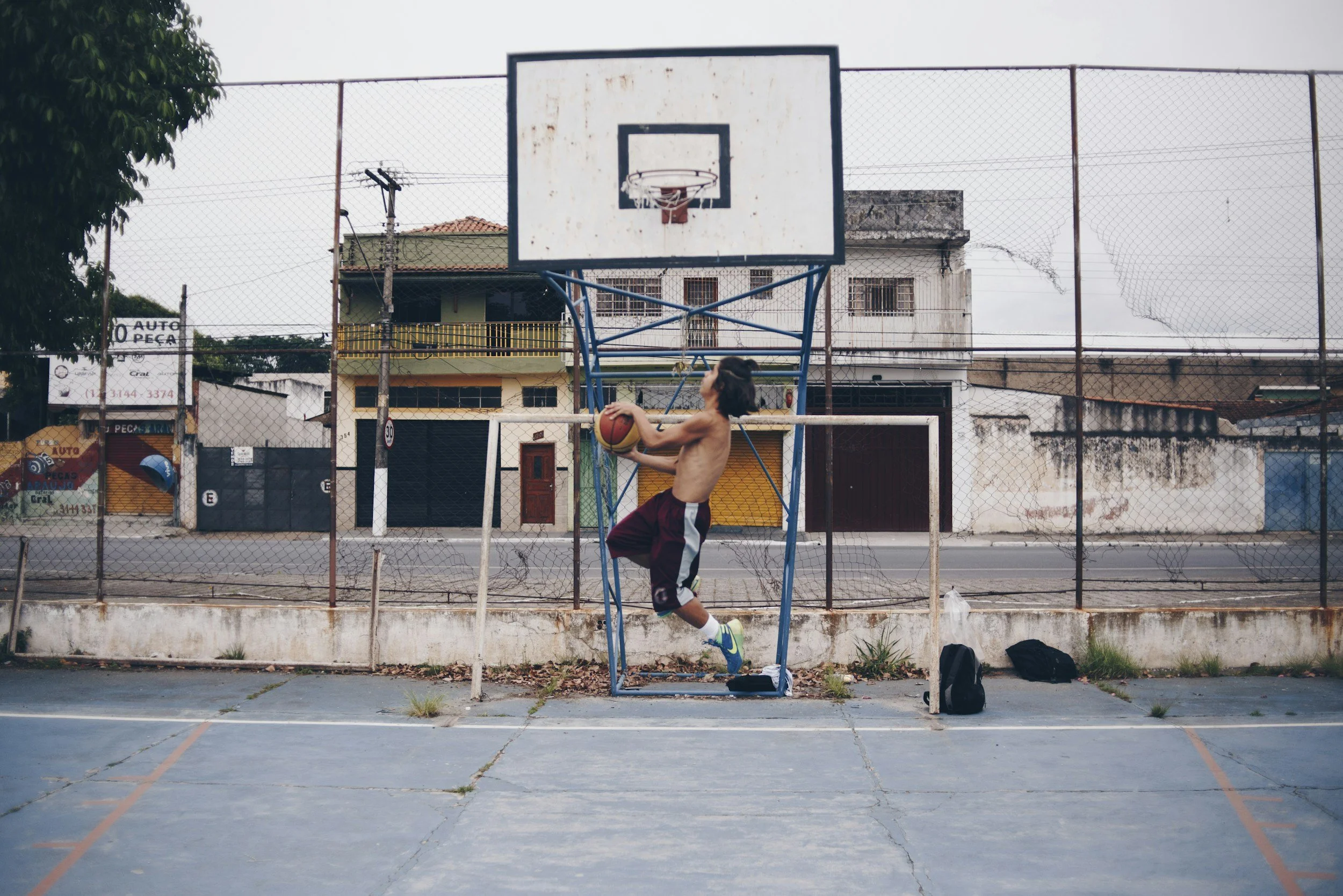 A shirtless young boy in basketball shorts and sneakers practicing basketball shots on an outdoor court, with a basketball hoop and a chain-link fence in the background.