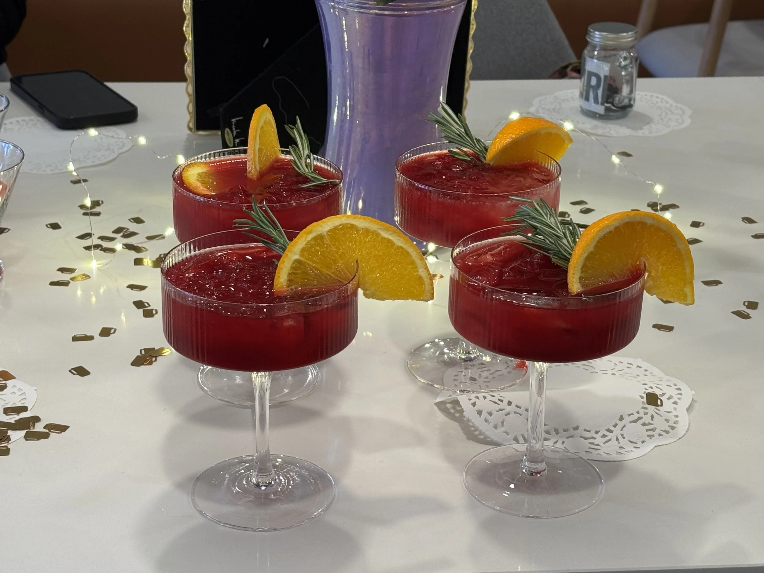 Four glasses of red cocktail garnished with orange slices and rosemary sprigs on a decorated table.
