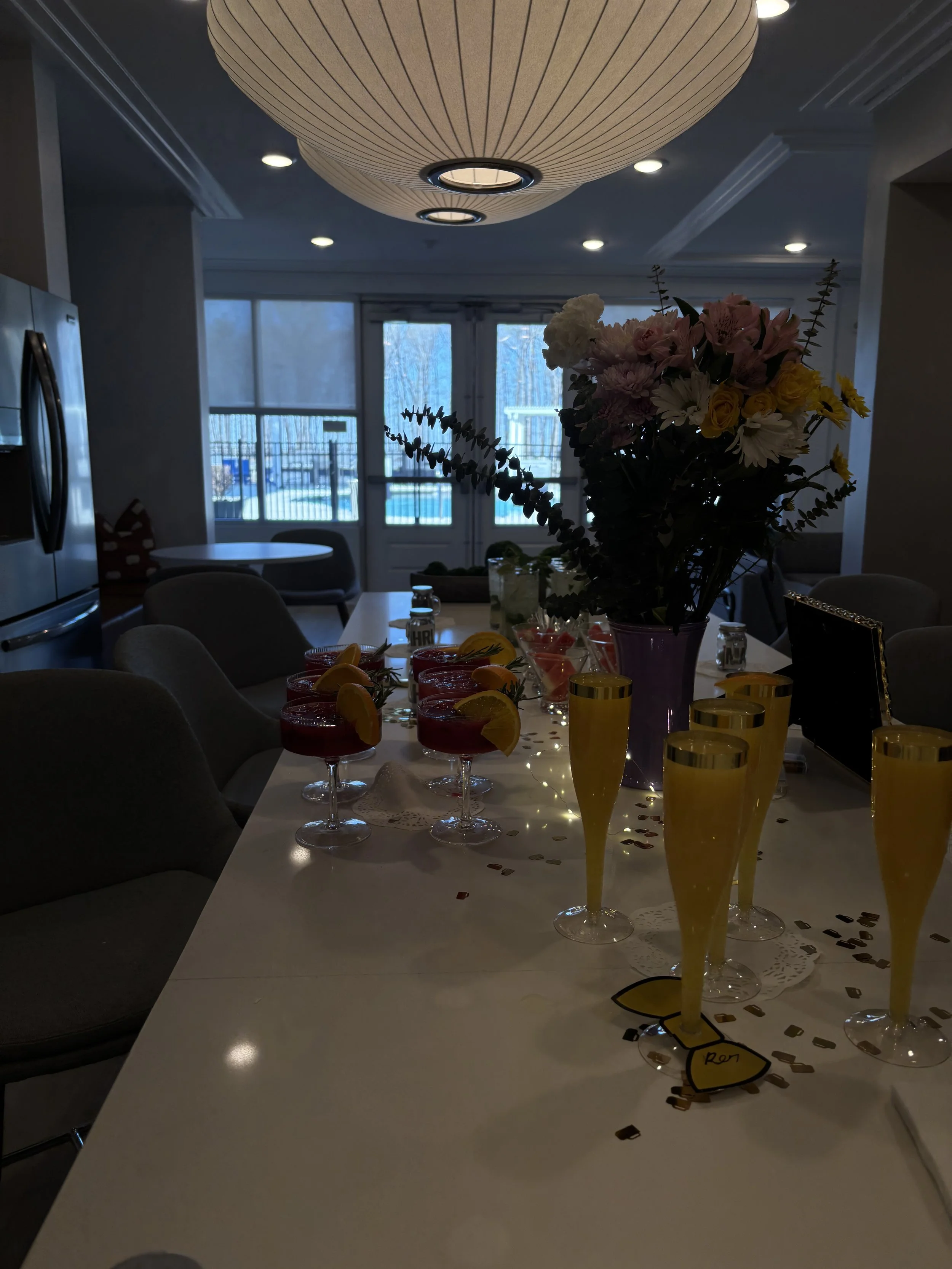 Party table with a large flower arrangement, glasses of drinks with lemon slices, and confetti, set in a bright indoor space with large windows and a view of a swimming pool outside.