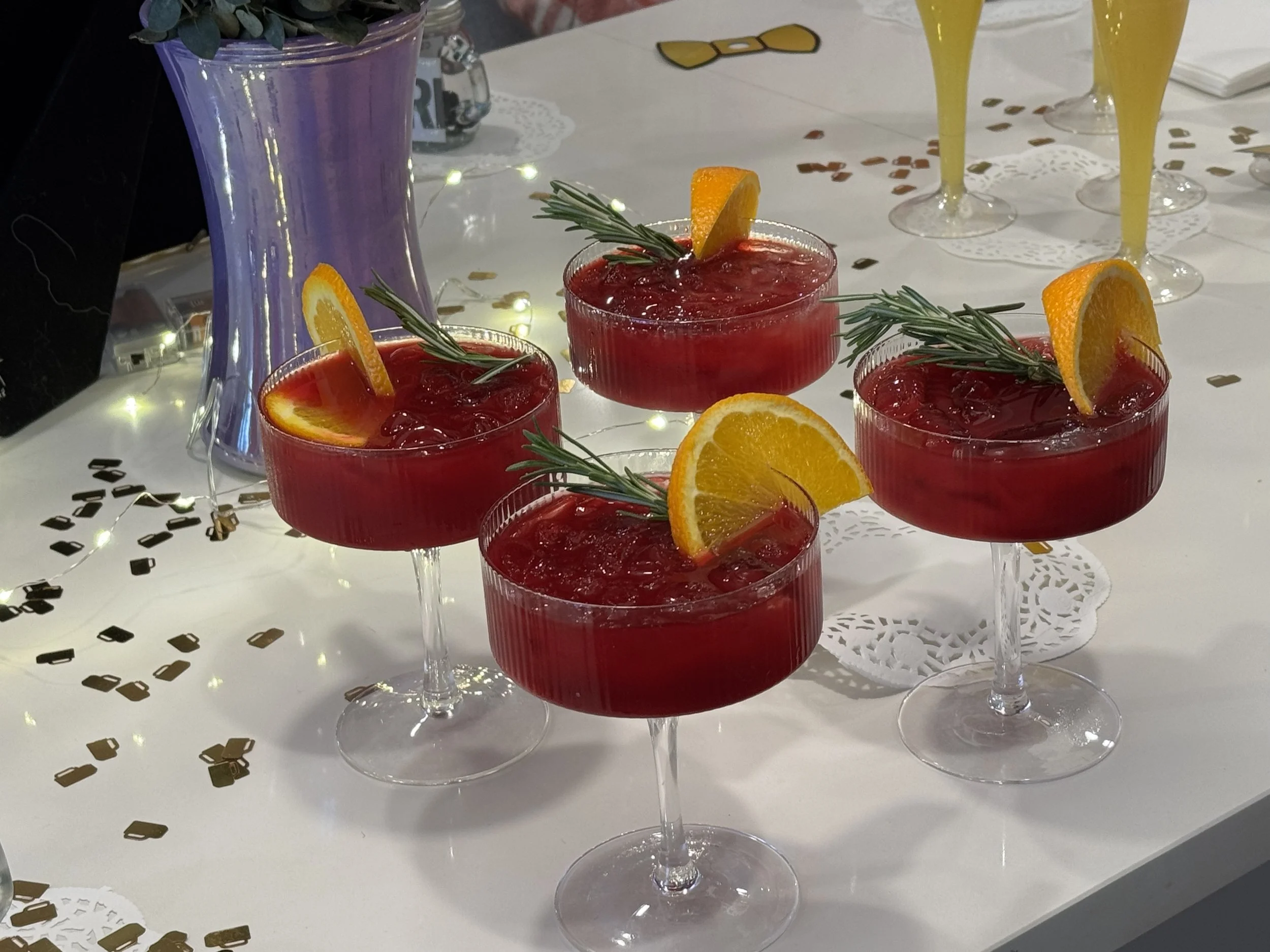 Four red cocktails garnished with orange slices and rosemary sprigs on a white table with confetti, a purple vase, and yellow drinks in the background.