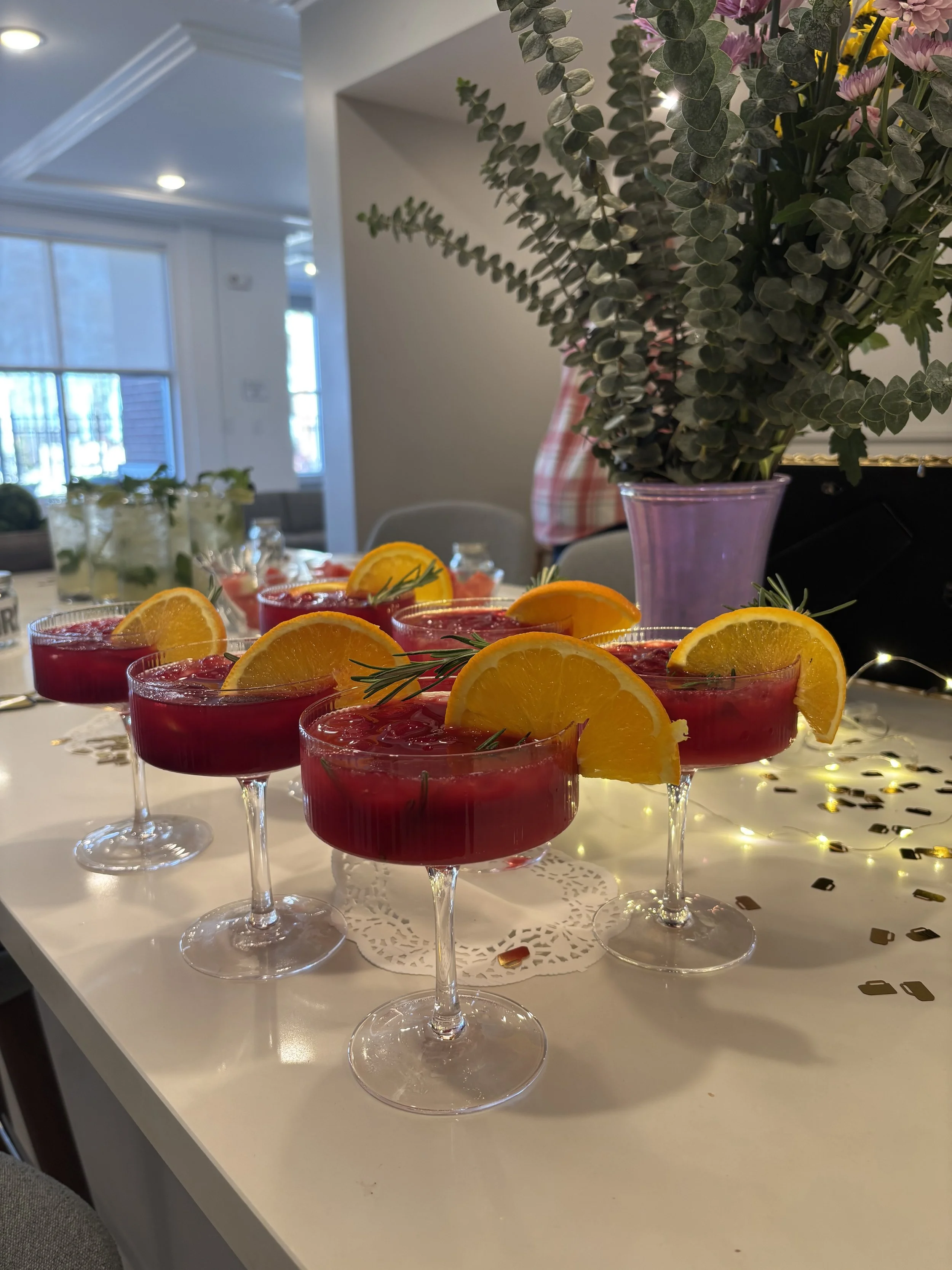 Six glasses of red cocktail with orange wedge and rosemary garnish on a white table, decorated with fairy lights and confetti, with a large vase of greenery and flowers in the background.