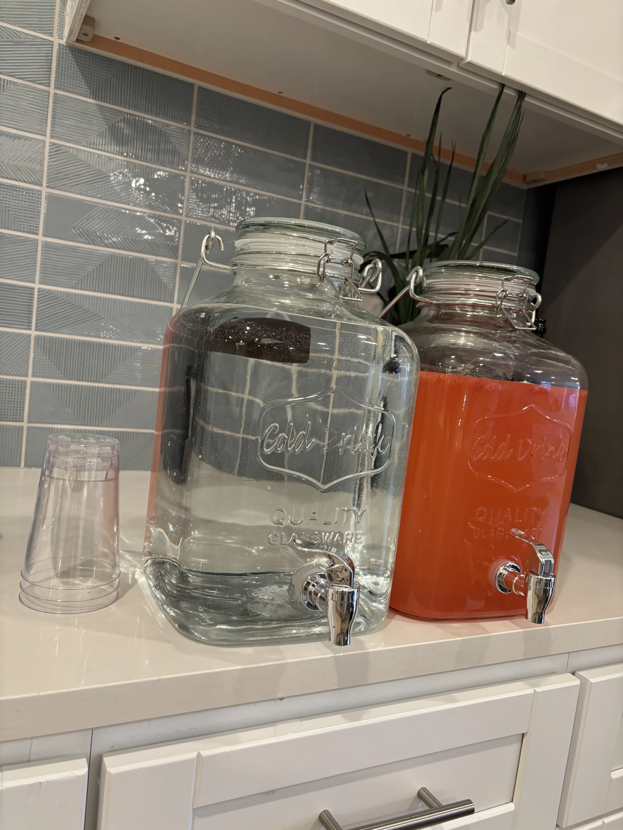 Two large glass beverage dispensers, one filled with water and the other with an orange-colored drink, with a small stack of clear plastic cups beside them, on a kitchen counter.