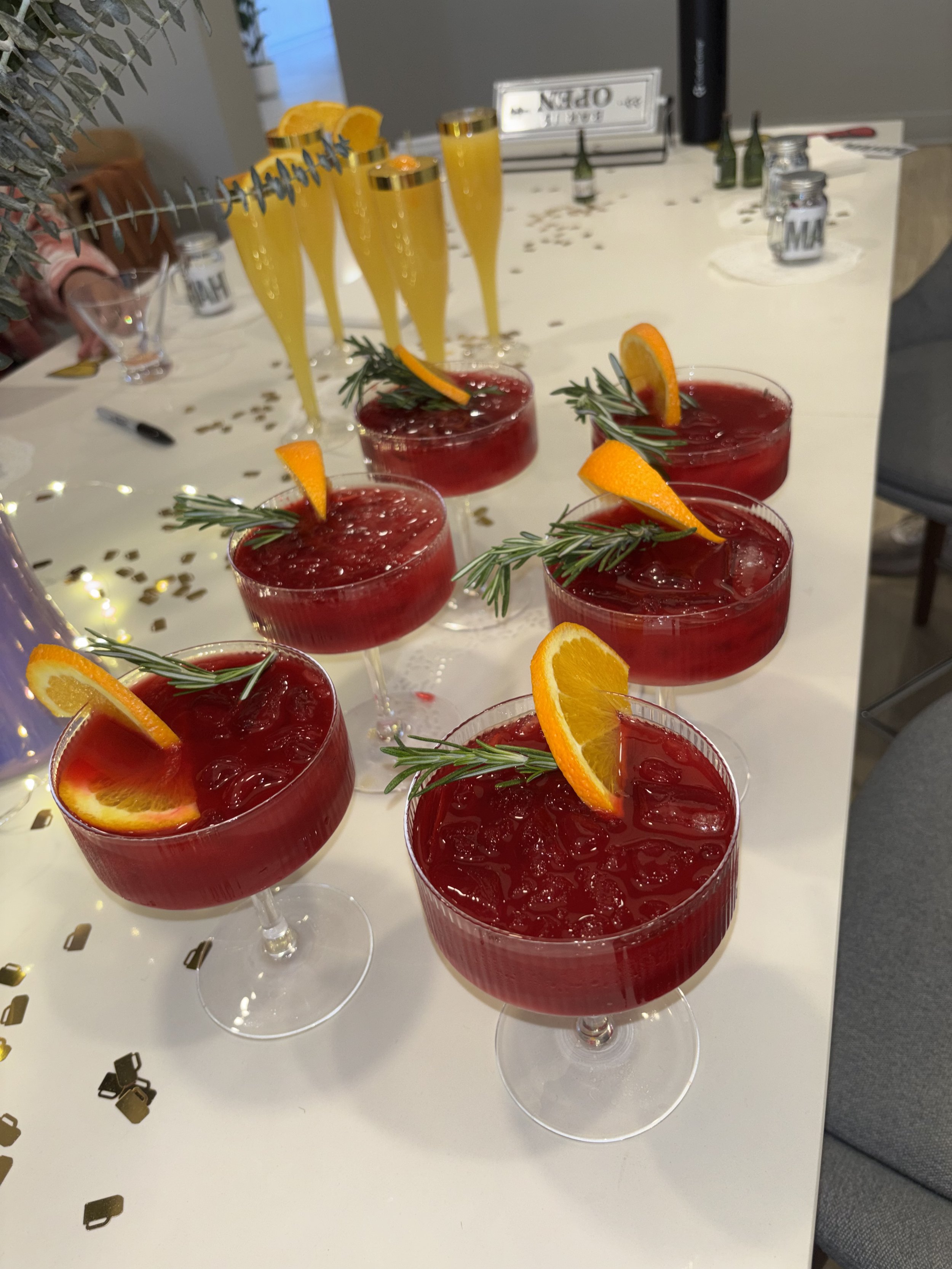 Six glasses of red punch garnished with orange slices and rosemary sprigs on a white table with gold confetti. In the background, several yellow drinks with orange slices and a sign that says 'OPEN' are visible.