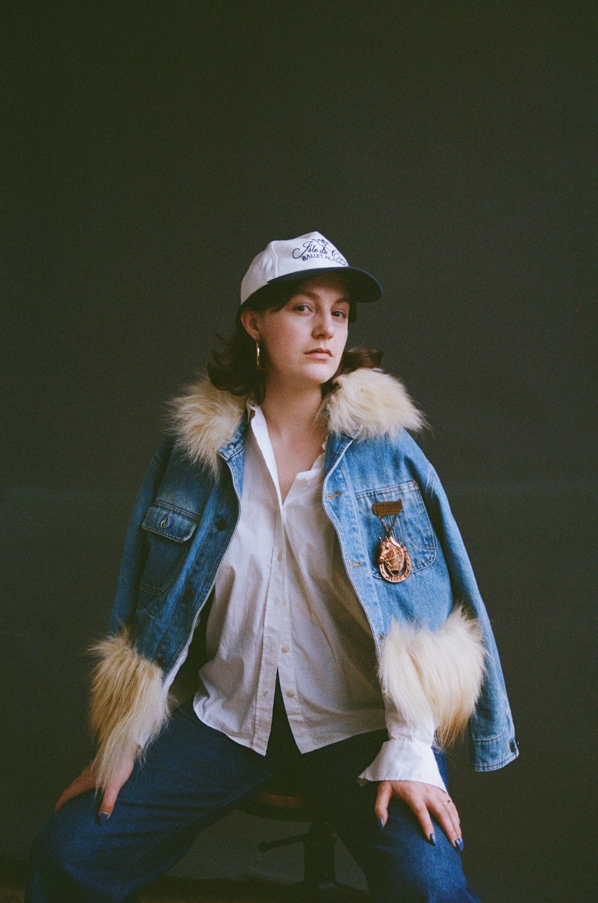 A woman with dark hair wearing a white cap, a blue denim jacket with a faux fur collar, a white shirt, and blue jeans, sitting on a black chair against a dark background.
