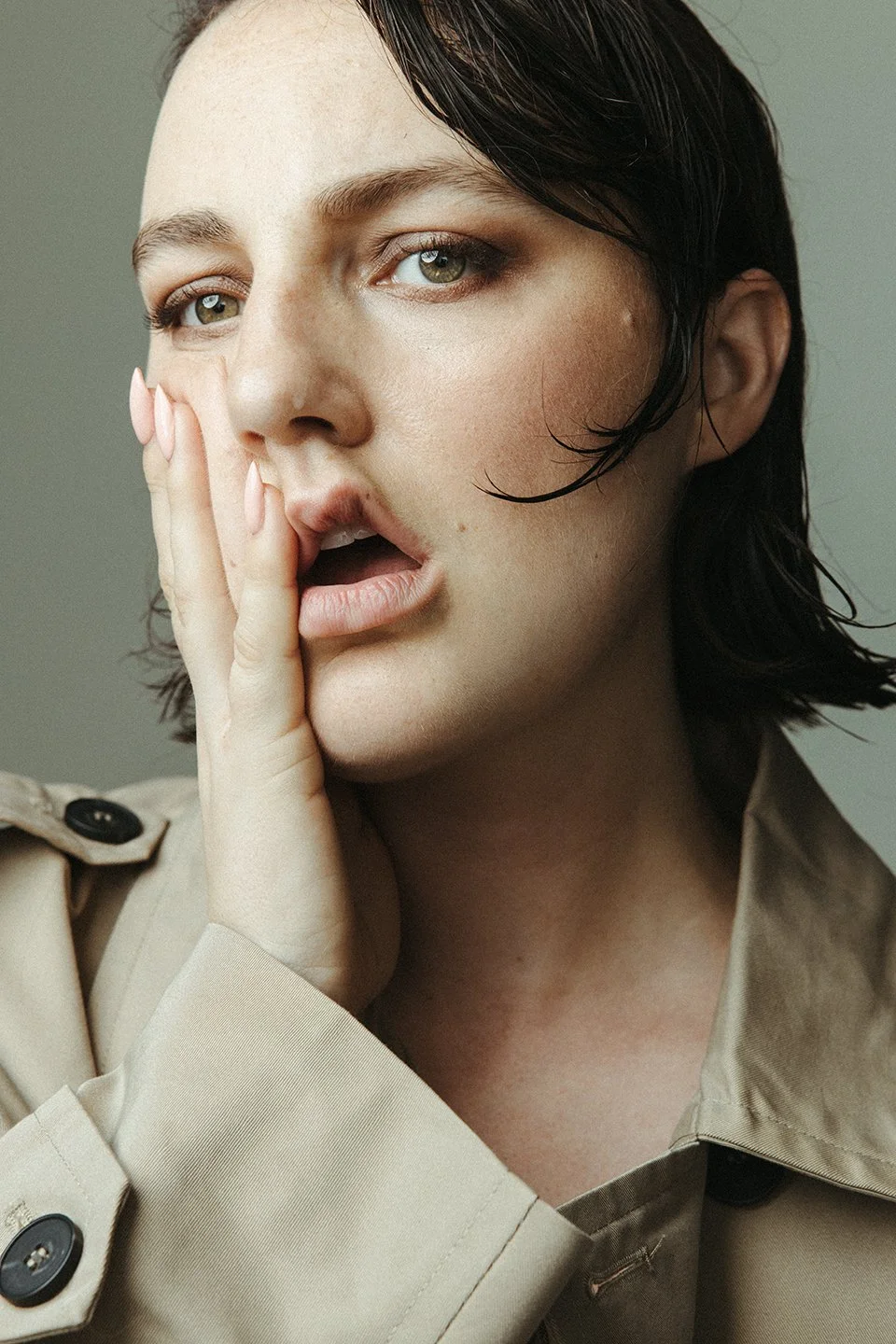 A close-up of a woman with short dark hair and green eyes touching her face with her hand while slightly opening her mouth, wearing a beige trench coat.