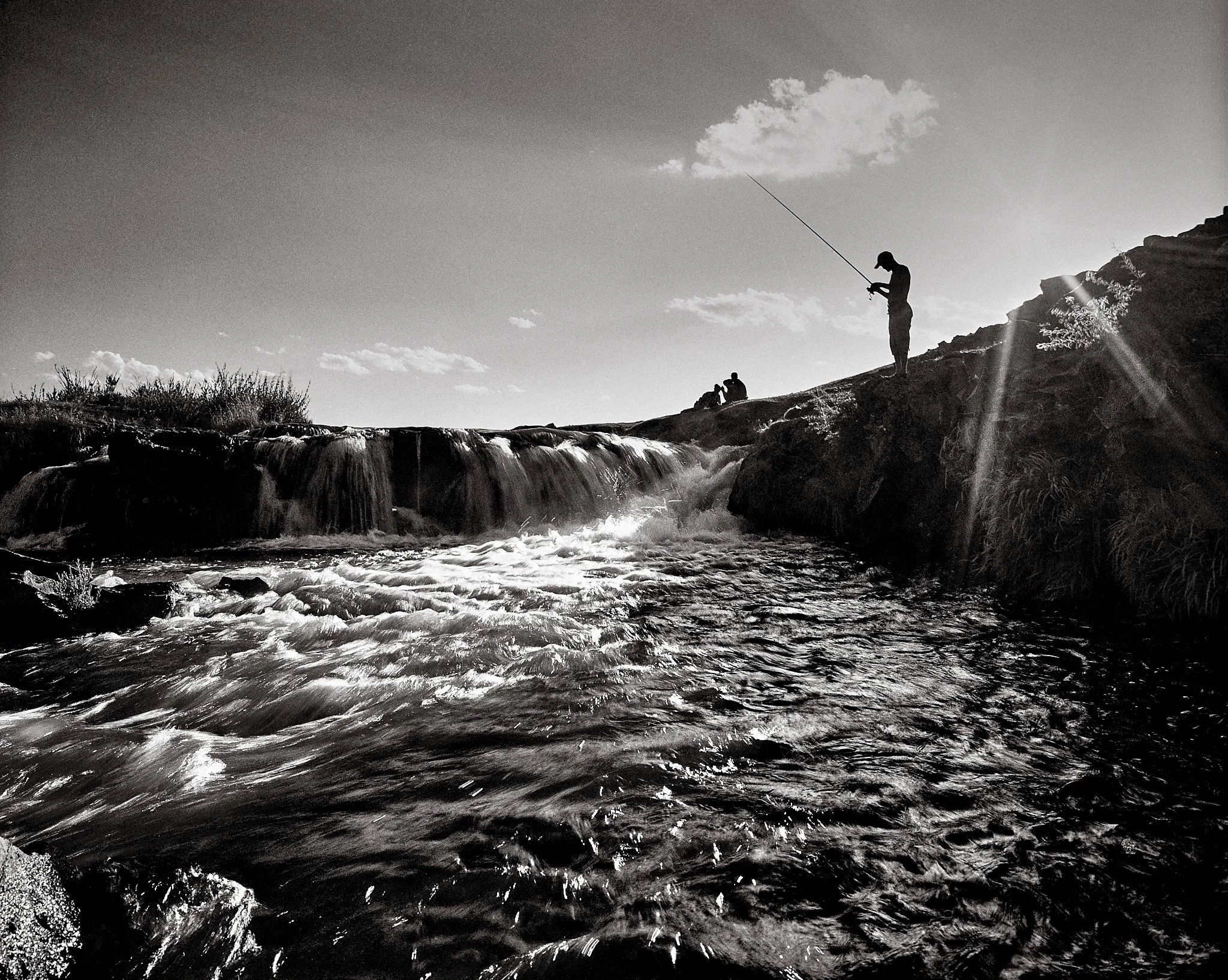 Silhouette of a person fishing by a small waterfall and flowing river in a natural outdoor setting, with a cloudy sky.
