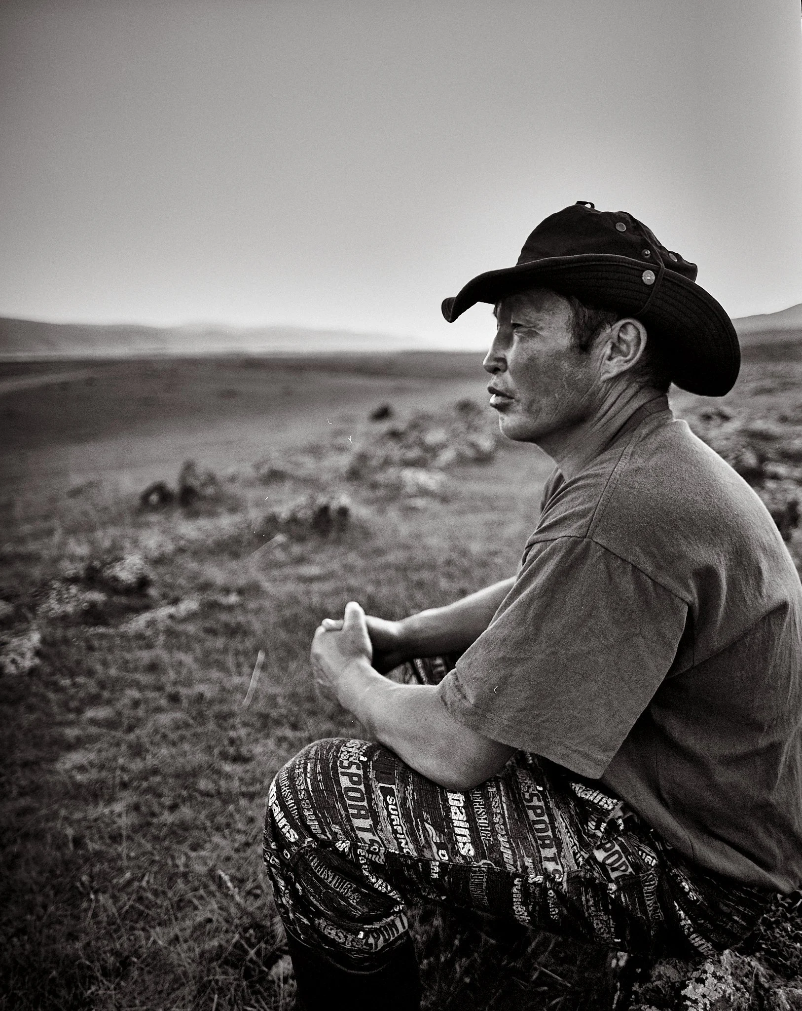 A man wearing a wide-brimmed hat and patterned pants sits cross-legged outdoors on a grassy field, looking into the distance with mountains in the background, captured in black and white.
