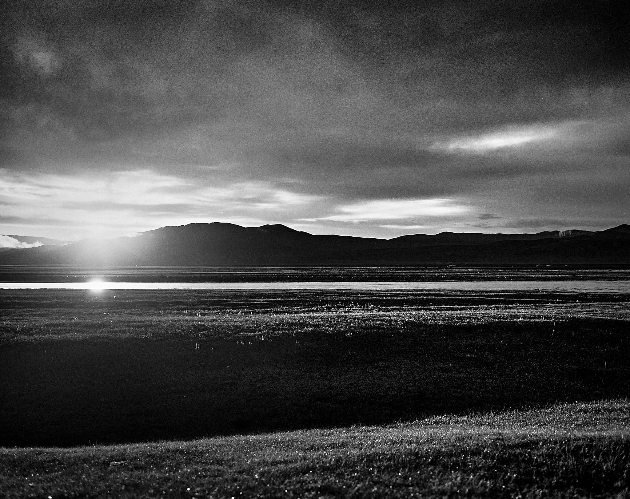 A black and white landscape featuring a mountain range in the distance, with the sun setting or rising near the horizon, casting reflections on a body of water, and a grassy foreground.