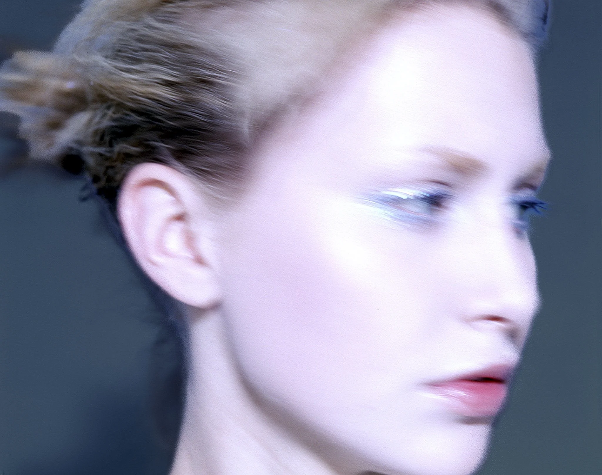 Close-up of a woman's face with her hair pulled back, wearing blue eyeshadow, looking to the side.