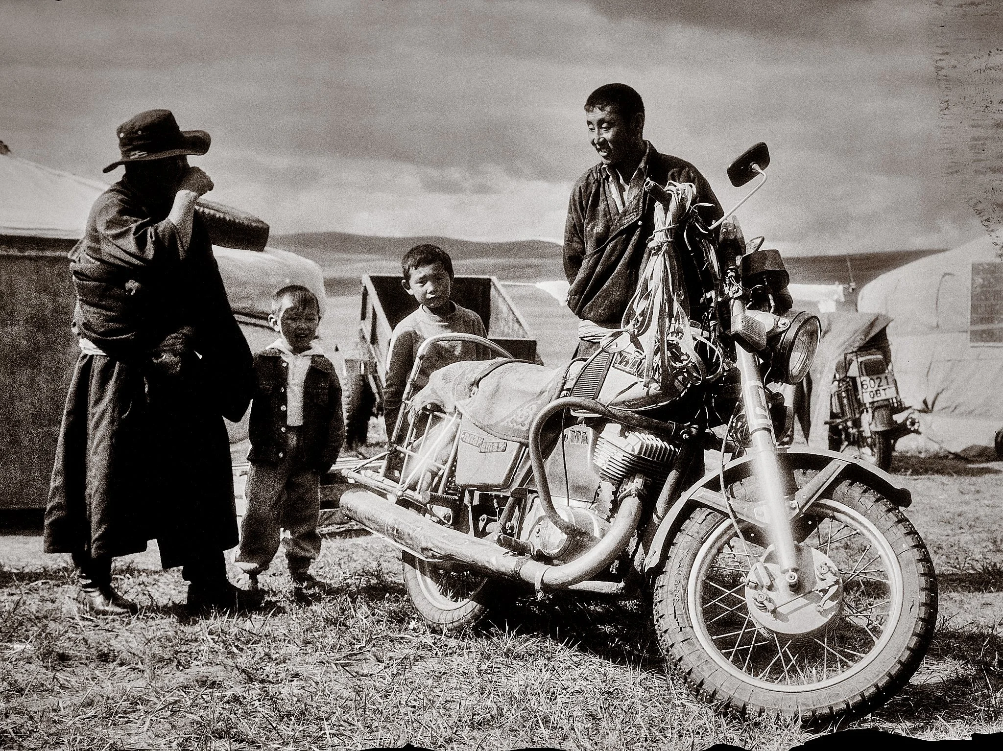 Black and white photo of five children and a motorcycle on grassy terrain, with tents and hills in the background.
