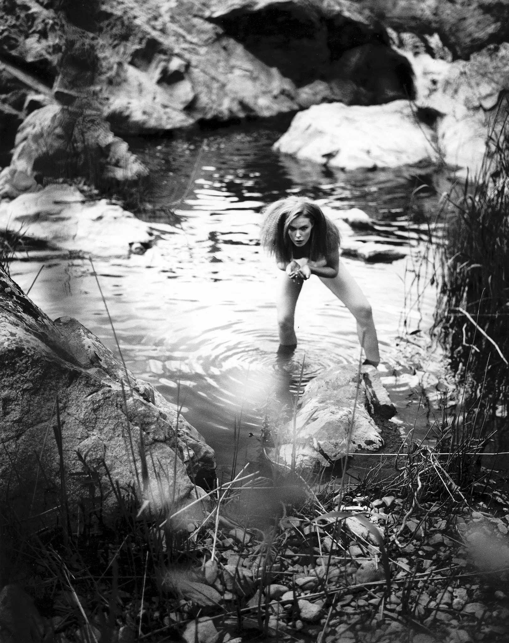 A woman with voluminous hair crouches in a rocky creek, looking intensely at the camera with her hands clasped in front of her. The scene is black and white, surrounded by rocks and tall grass.