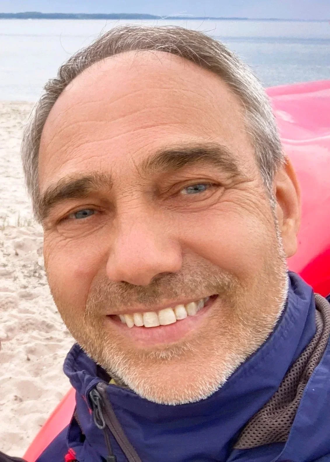 Nahaufnahme eines lachenden Mannes mit grauem Haar und blauen Augen am Strand, im Hintergrund Wasser und Sand, trägt eine blaue Jacke.