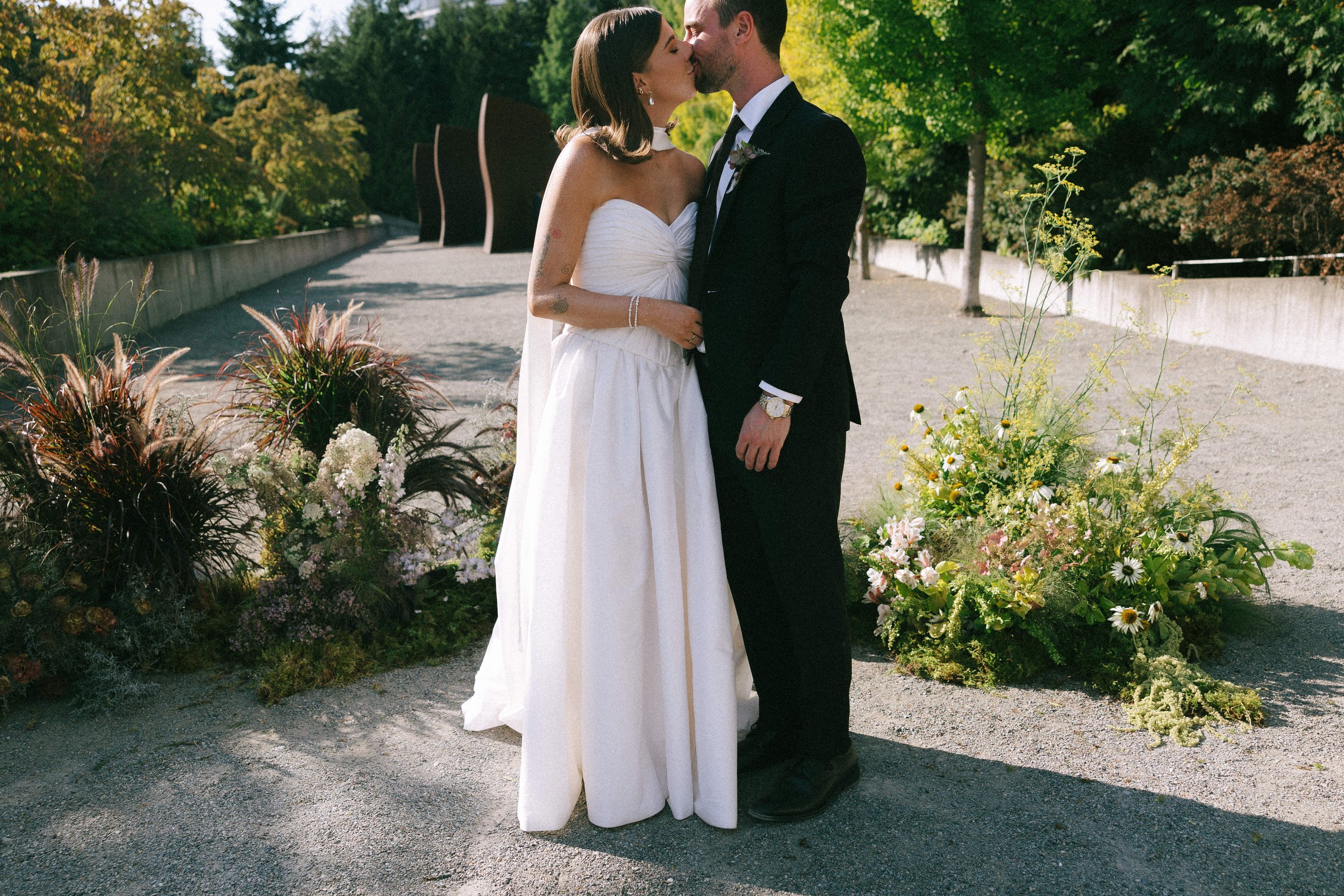 A newlywed couple sharing a kiss outdoors on a sunny day, with floral arrangements and greenery in the background.