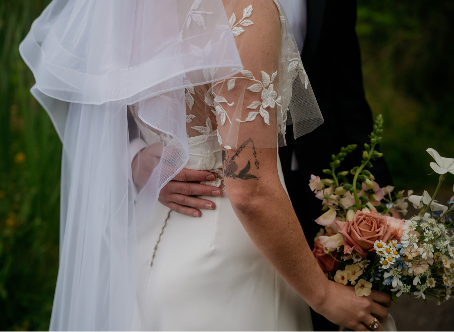 Close-up of a bride and groom on their wedding day, with the bride wearing a white gown and a sheer, embroidered veil, holding a bouquet of pink, white, and cream flowers, and the groom dressed in a black suit.