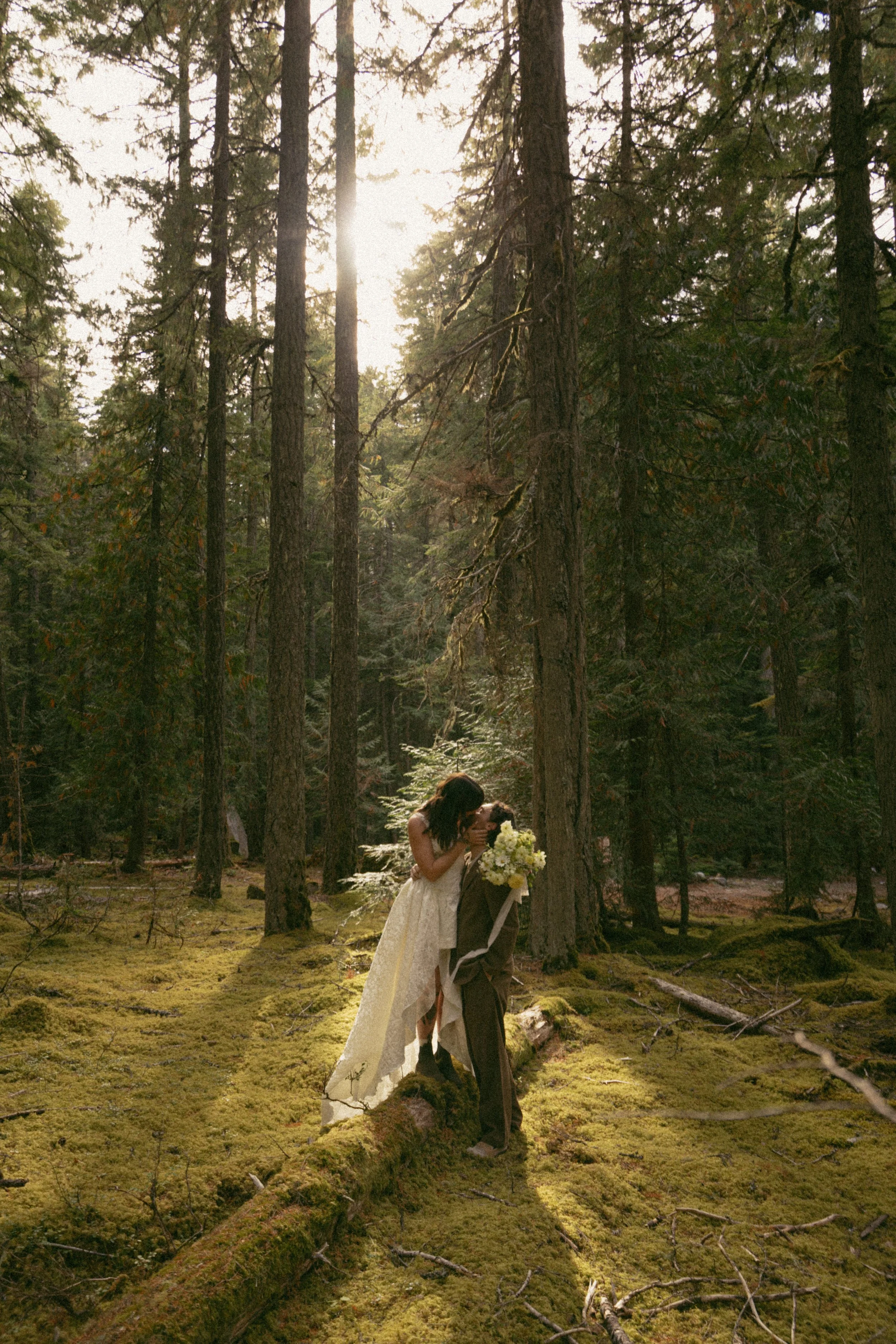 A couple dressed in wedding attire, the bride in a white dress and the groom in a suit, in a forest with tall trees and sunlight filtering through the trees.