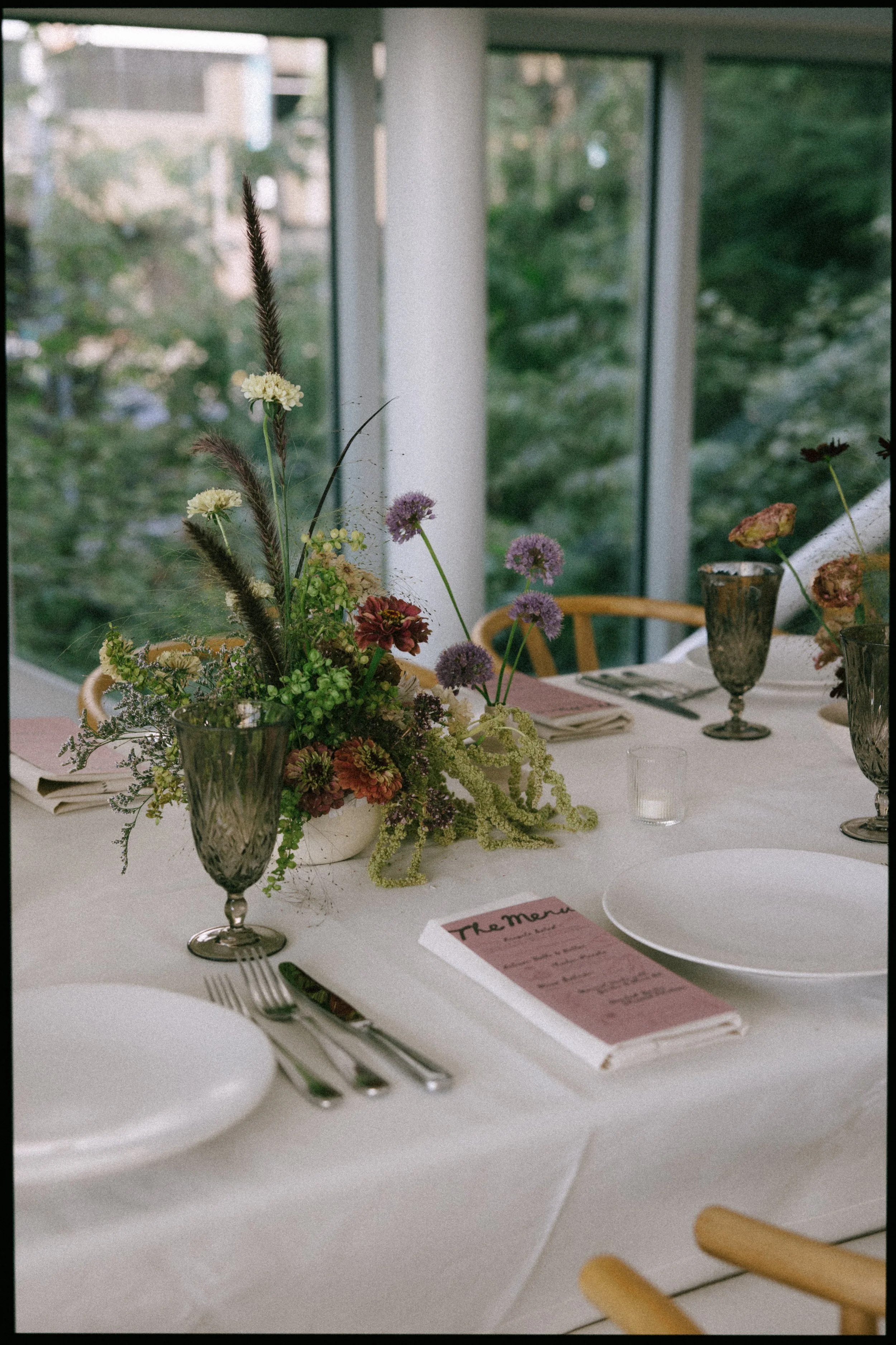 A table set for a meal with a floral centerpiece, white plates, silverware, purple and pink glassware, and a pink menu card, in front of large windows showing greenery outside.