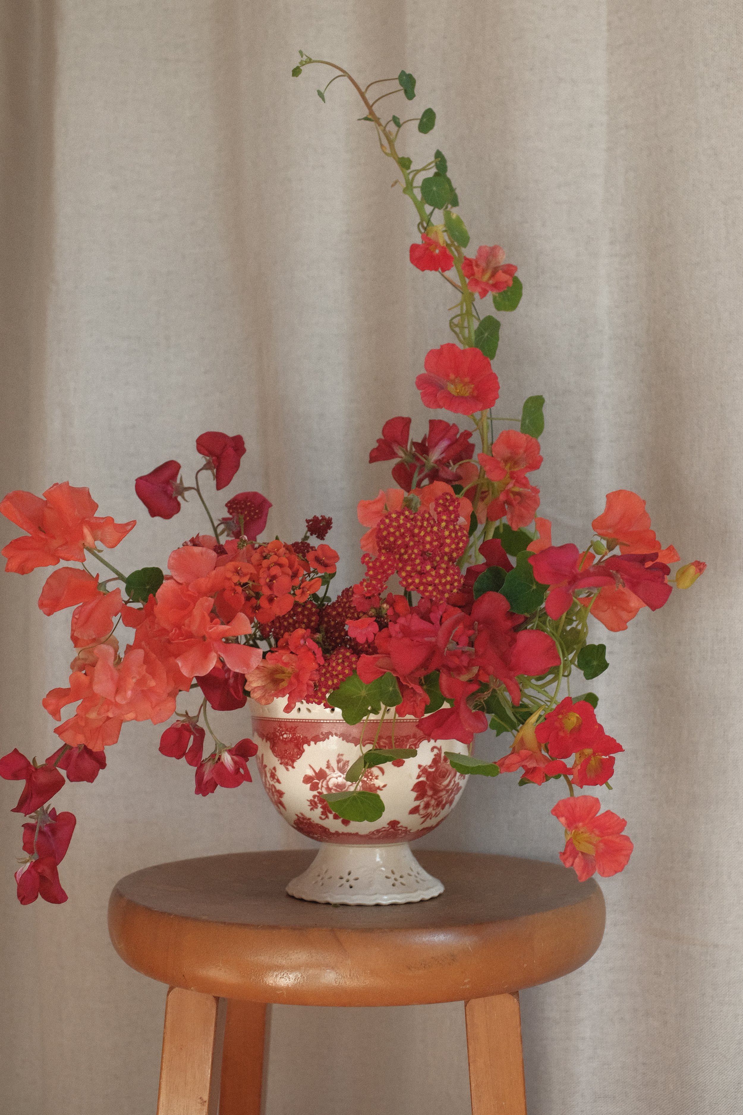 A floral arrangement in a red and white decorative vase placed on a wooden stool against a beige curtain background.