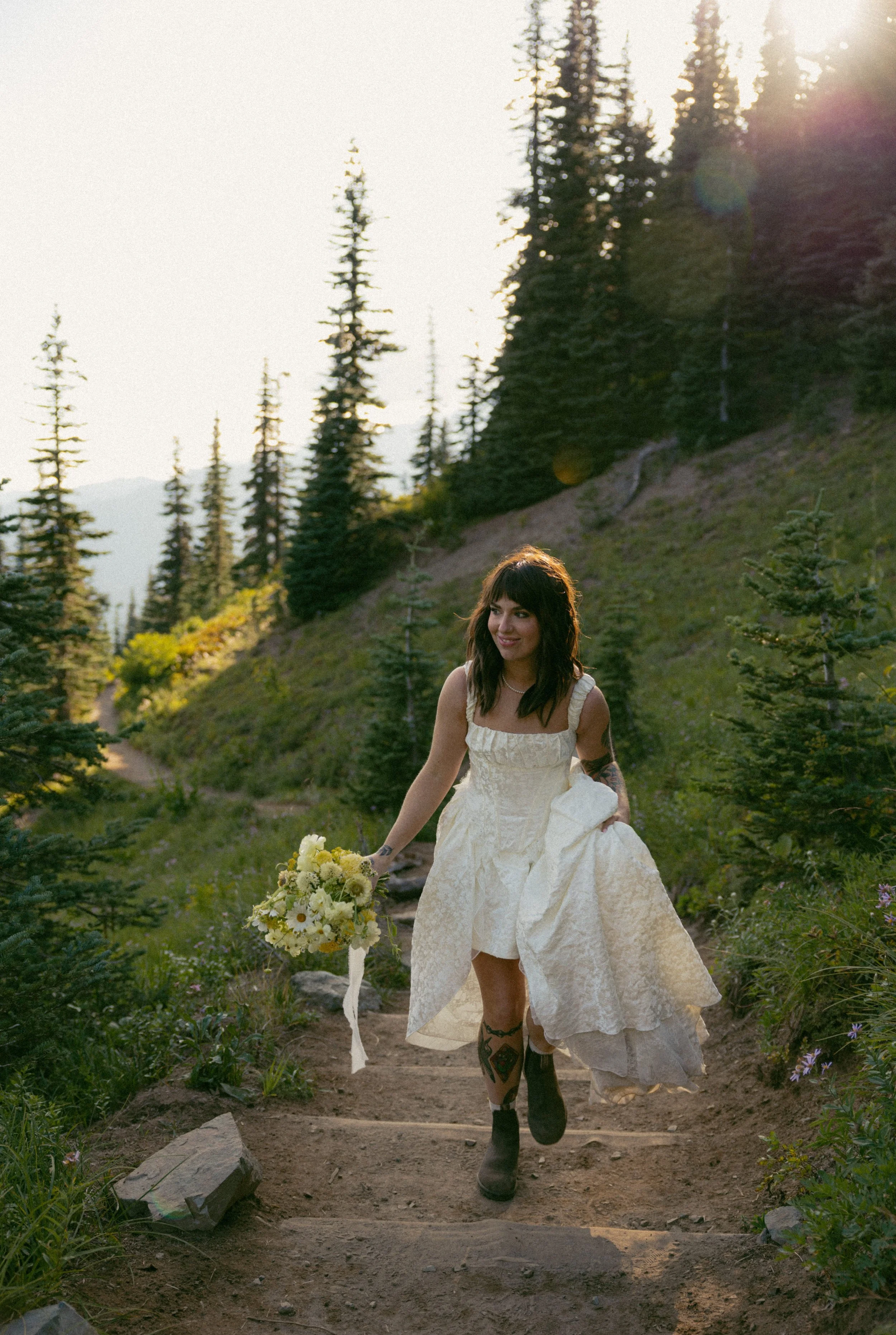 A woman in a white lace dress holding a bouquet of flowers walking down outdoor steps in a forested area during sunset.