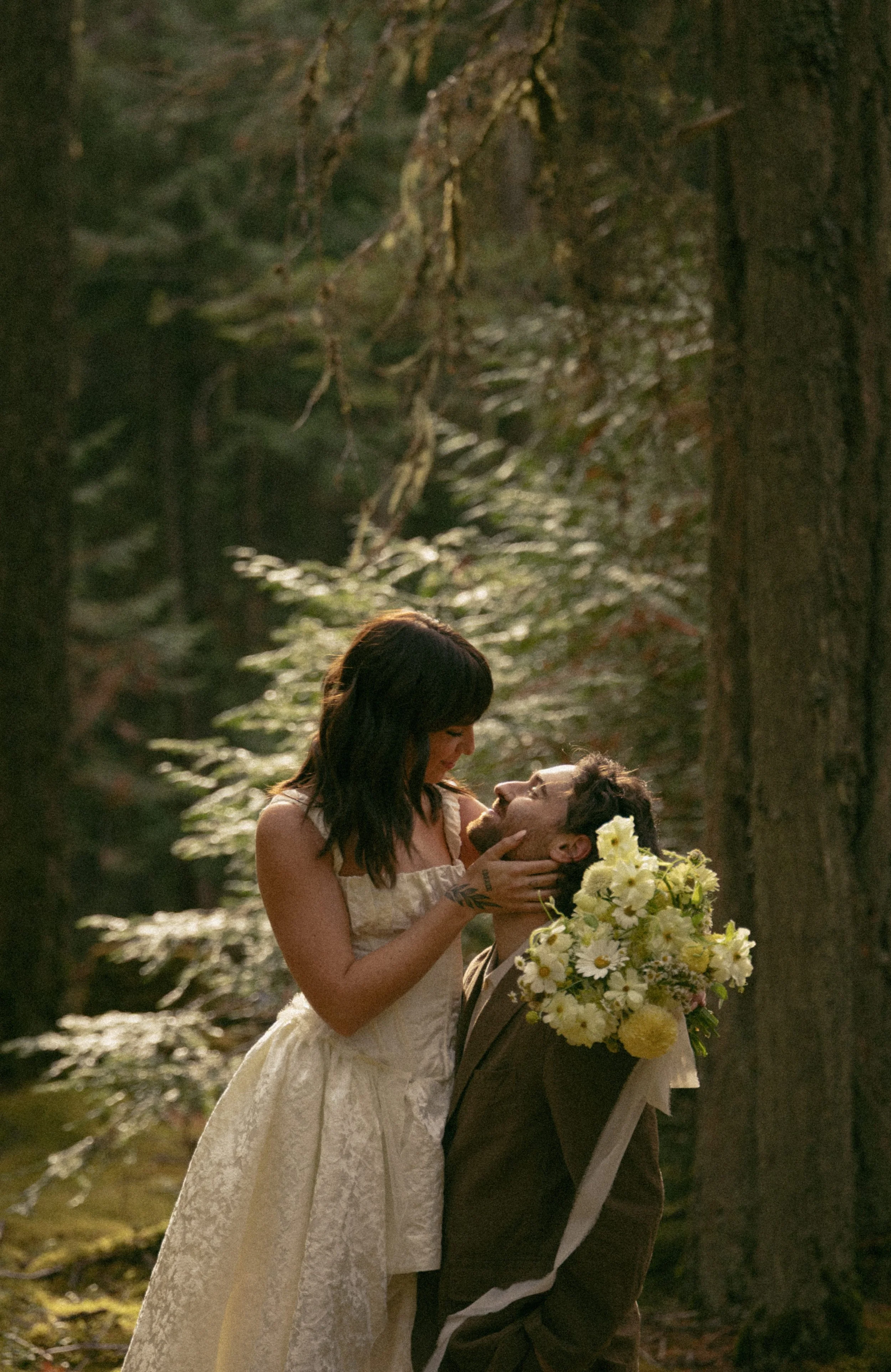 A woman and a man in a forest, with the woman holding the man's face and the man holding a bouquet of white flowers.