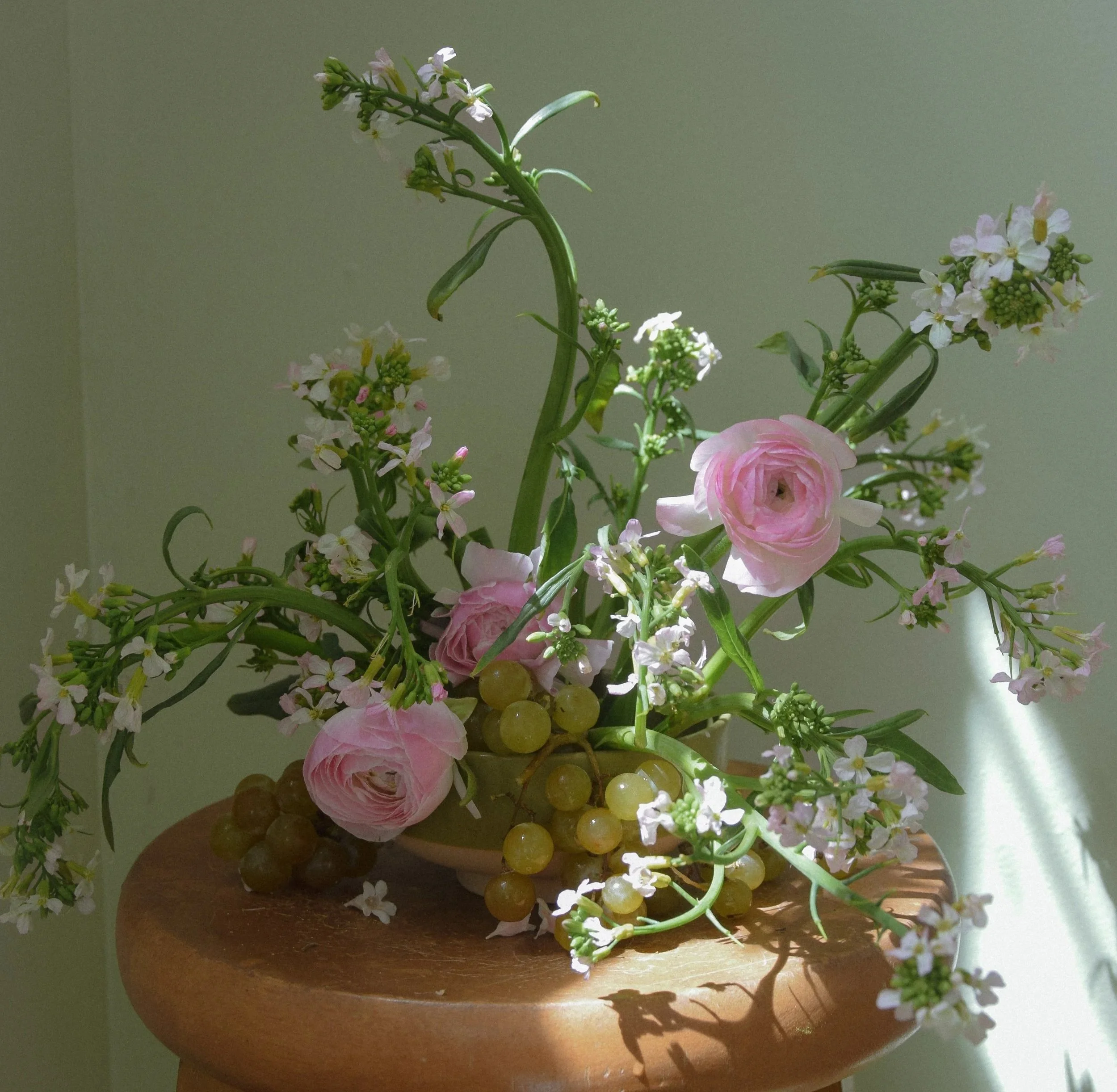 A floral arrangement with pink roses, small white flowers, green grape clusters, in a white bowl on a wooden surface, with soft natural light.