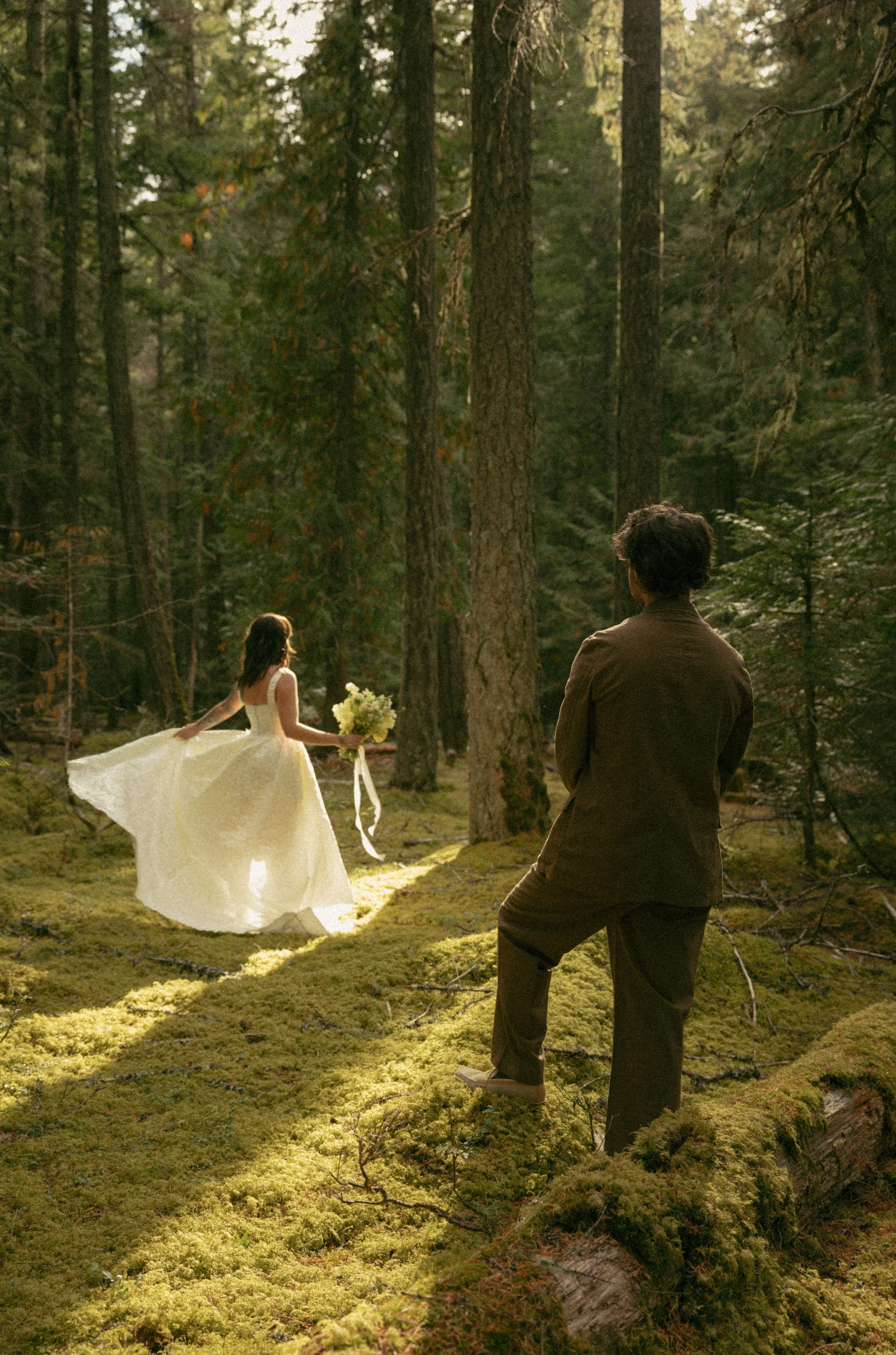 A woman in a white dress holding a bouquet walking in a forest, while a man in dark clothing watches her from a fallen log.