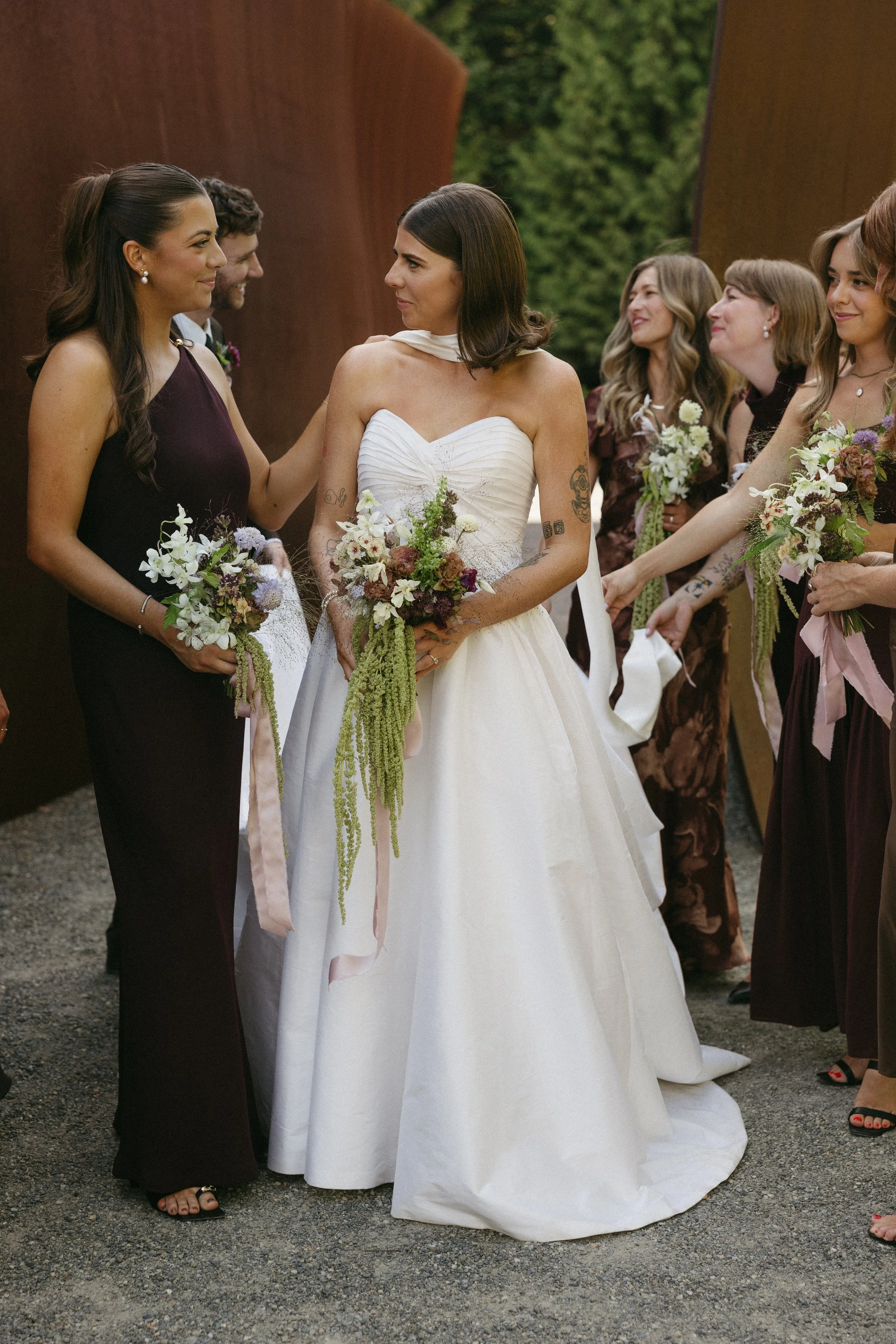 A group of women in formal dresses, with one woman in a white wedding gown, holding bouquets, standing outdoors and interacting during a wedding ceremony.