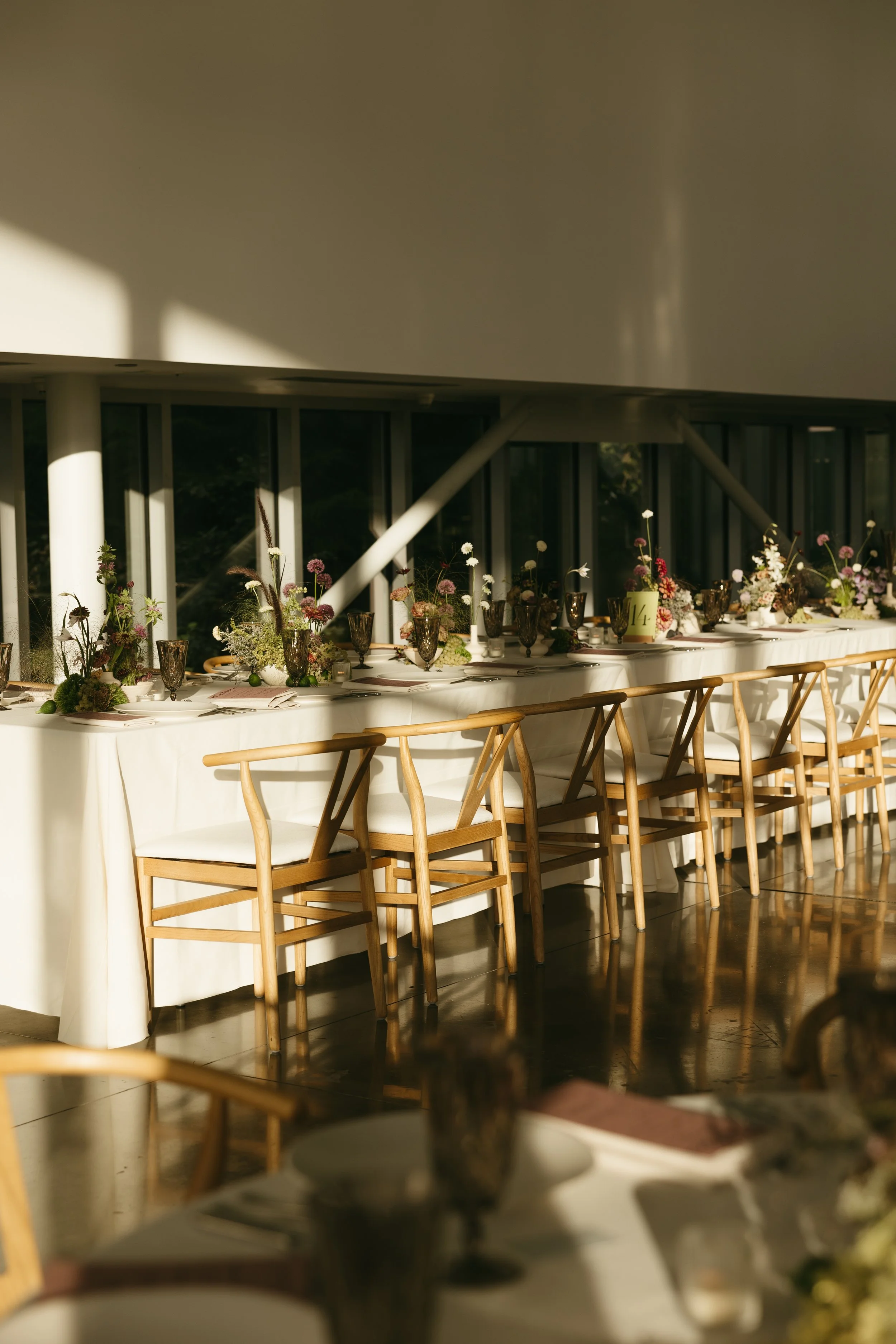 Long banquet table set for a formal event with elegant glassware, plates, napkins, and floral centerpieces, with sunlight streaming through large windows.