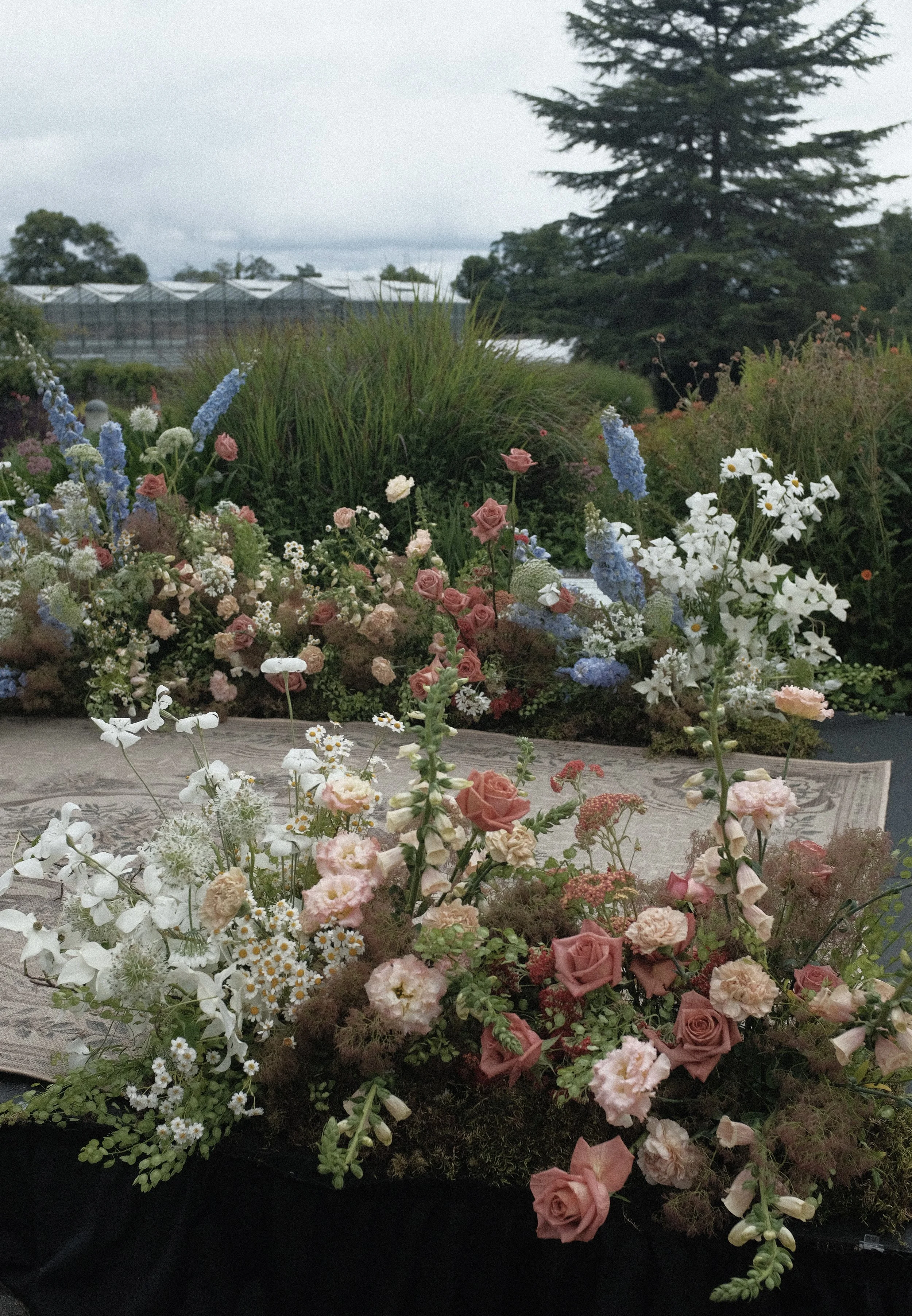 A colorful arrangement of assorted flowers, including roses and daisies, placed on a table outdoors with a lush garden and trees in the background.