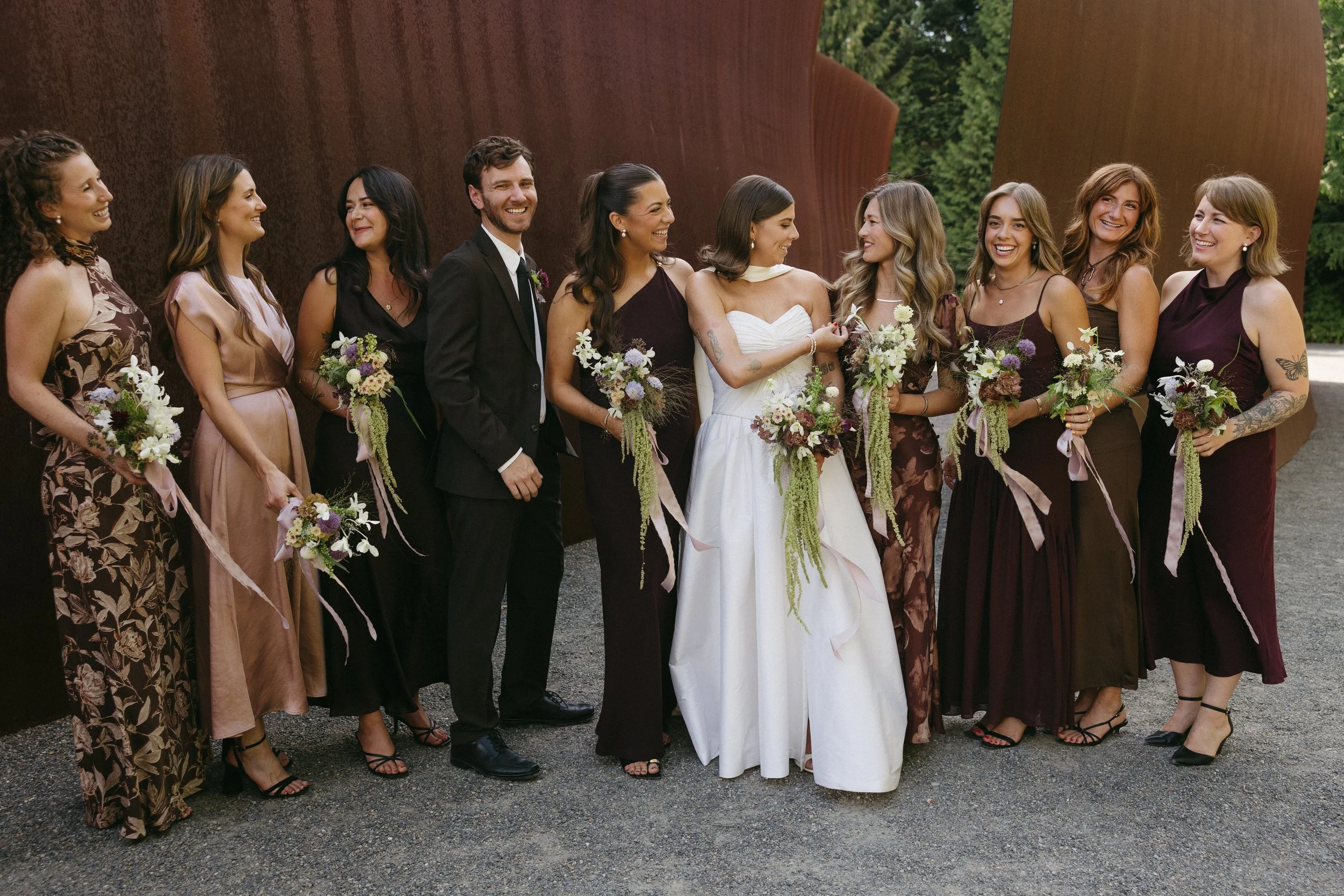 A group of twelve people, including one man and eleven women, standing in a line outdoors, dressed formally for a wedding, with some holding bouquets of flowers. The scenery includes a rusty metal wall and green trees in the background.