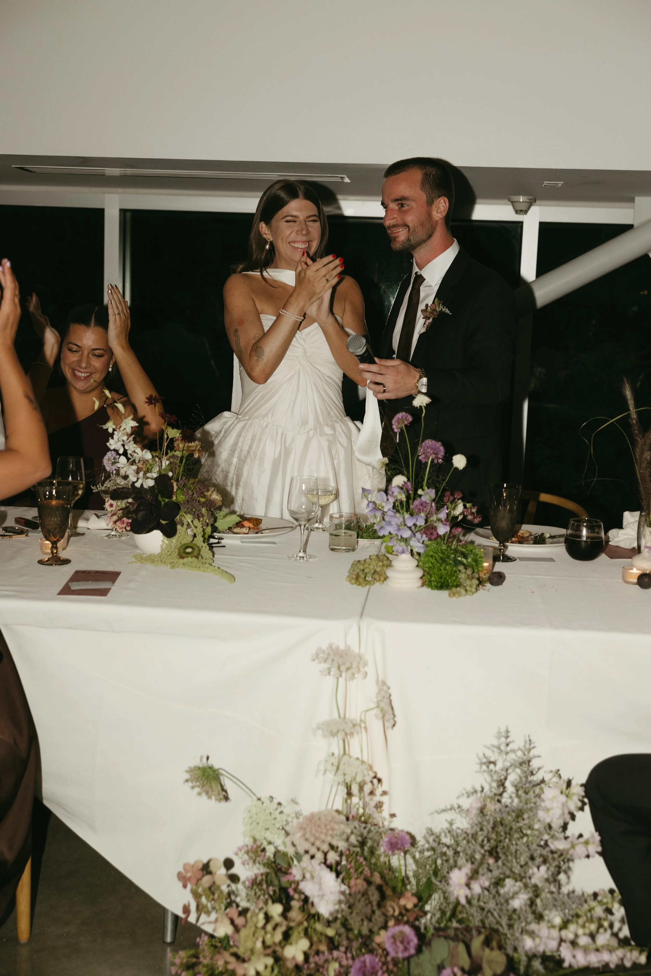 A bride and groom stand at a wedding reception table, smiling and laughing. The bride wears a white dress and appears emotional, while the groom in a black suit holds a microphone. Guests are seated around the table with floral arrangements and wine 