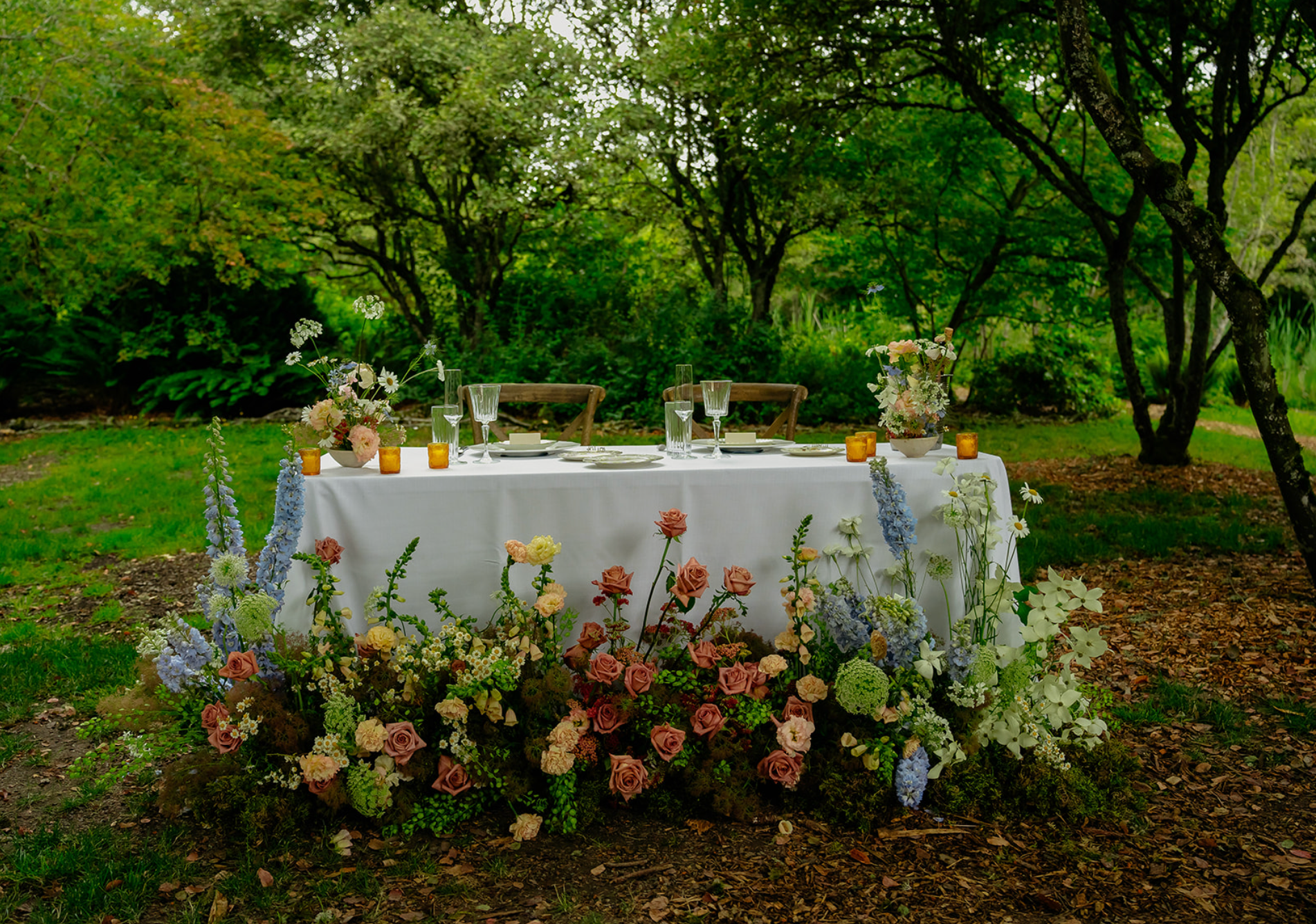 An outdoor table set for a formal event in a lush, green garden with trees. The table is covered with a white cloth and decorated with various flower arrangements in pastel colors, candles, plates, glasses, and silverware.