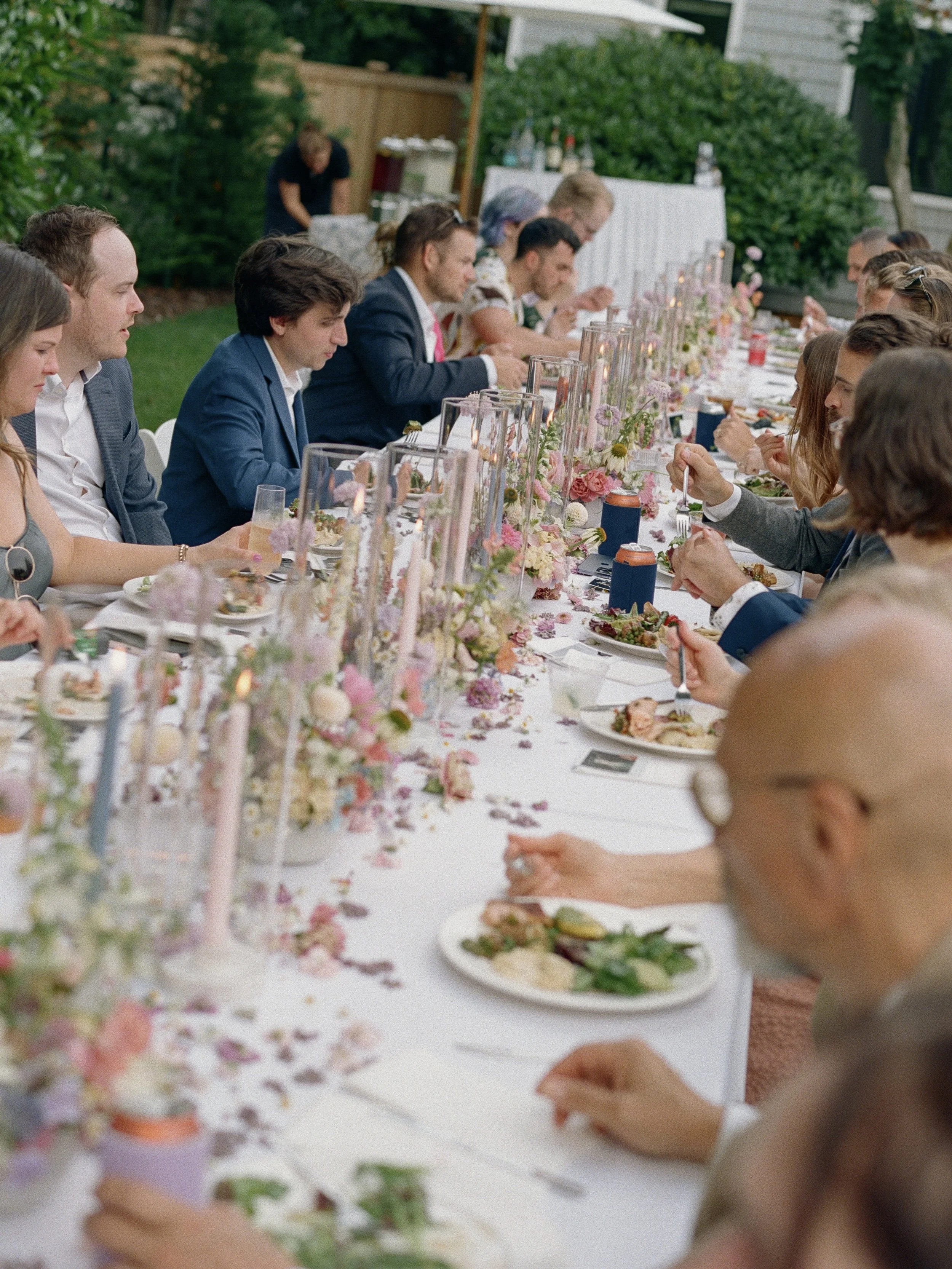 People sitting at a long outdoor banquet table decorated with flowers and candles.